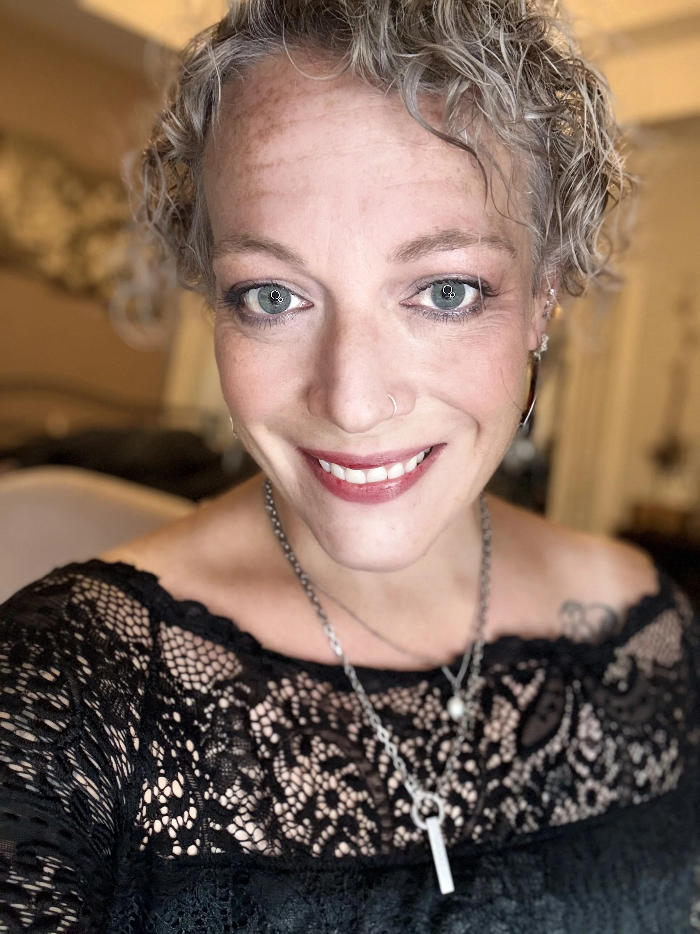 A woman with curly gray hair smiling at the camera, wearing a black lace top and a necklace with a pendant.