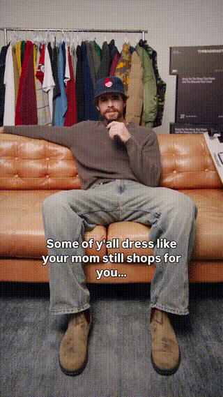 A man sitting on a sofa with a relaxed posture, wearing a baseball cap, sweatshirt, and jeans, in front of a clothing rack filled with colorful shirts and jackets, with a television behind him. Subtitles read: "Some of y'all dress like your mom still shops for you..."