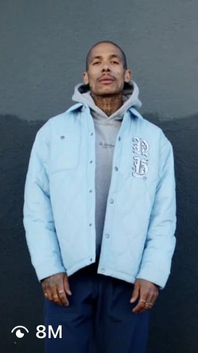 Person standing outdoors against a dark background, wearing a light blue jacket and hoodie, with a tattoo on neck, looking at the camera.