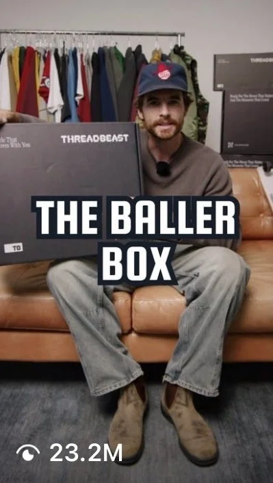 A man sitting on a brown leather sofa holding a black box with the label 'THE BALLER BOX' in what appears to be a clothing store or office with clothes hanging on a rack behind him.