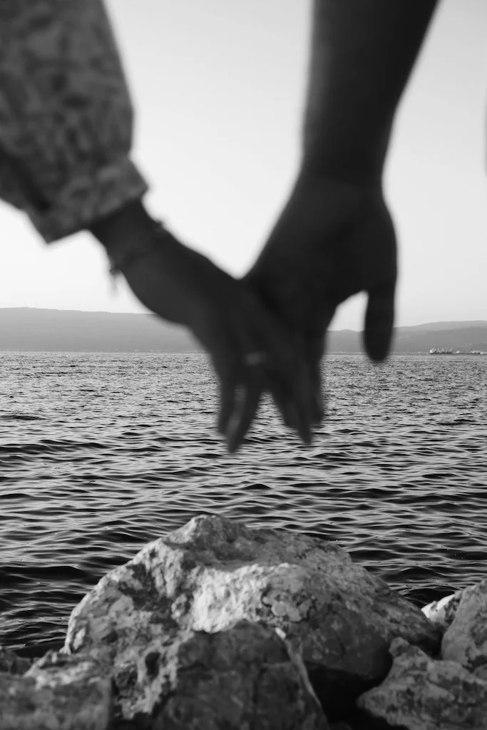 Two people holding hands by the water with rocks in the foreground and a distant shoreline in the background.