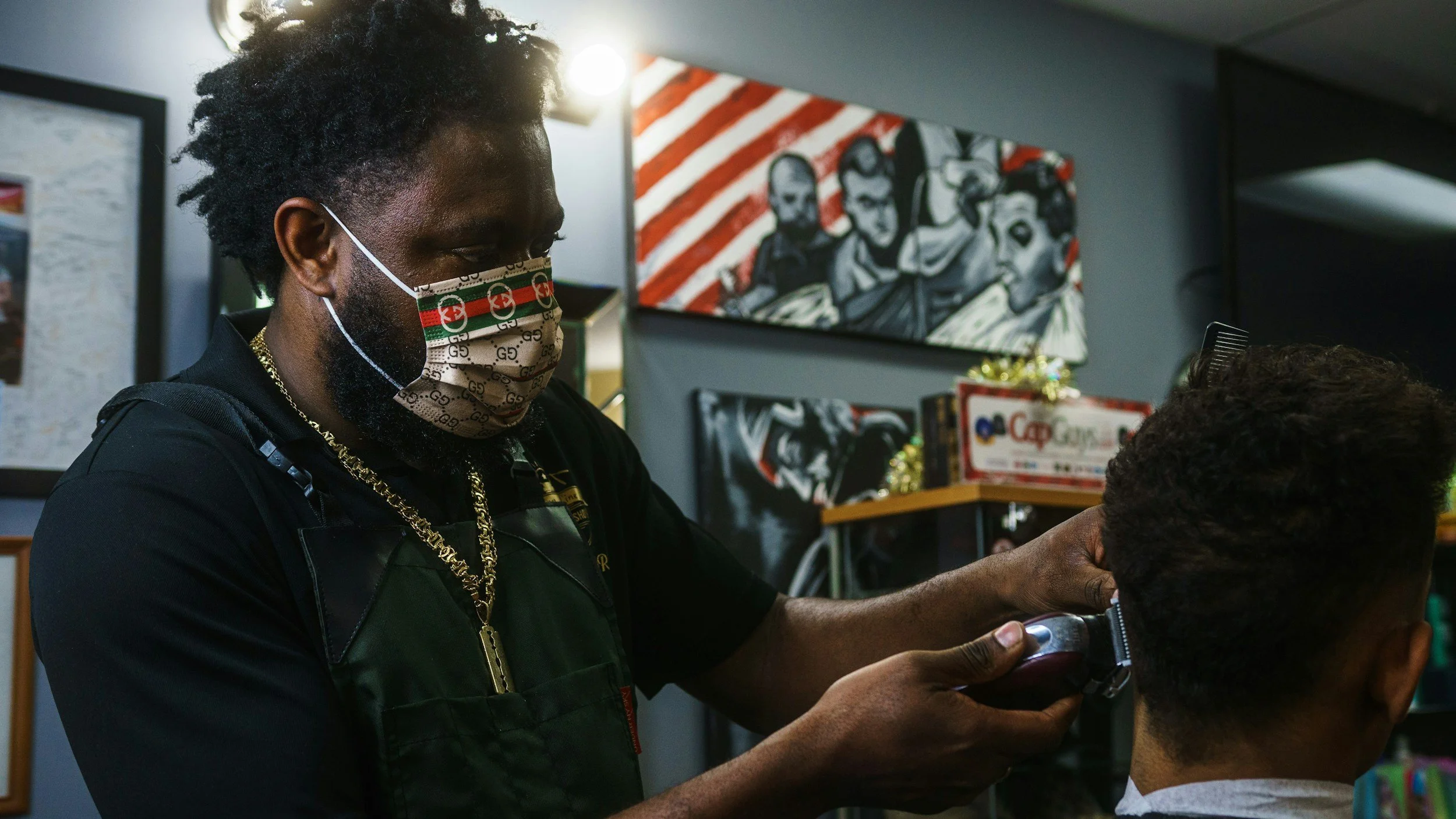 Barber wearing a Gucci face mask and gold chains is trimming a customer's hair in a barbershop decorated with artwork and framed posters.
