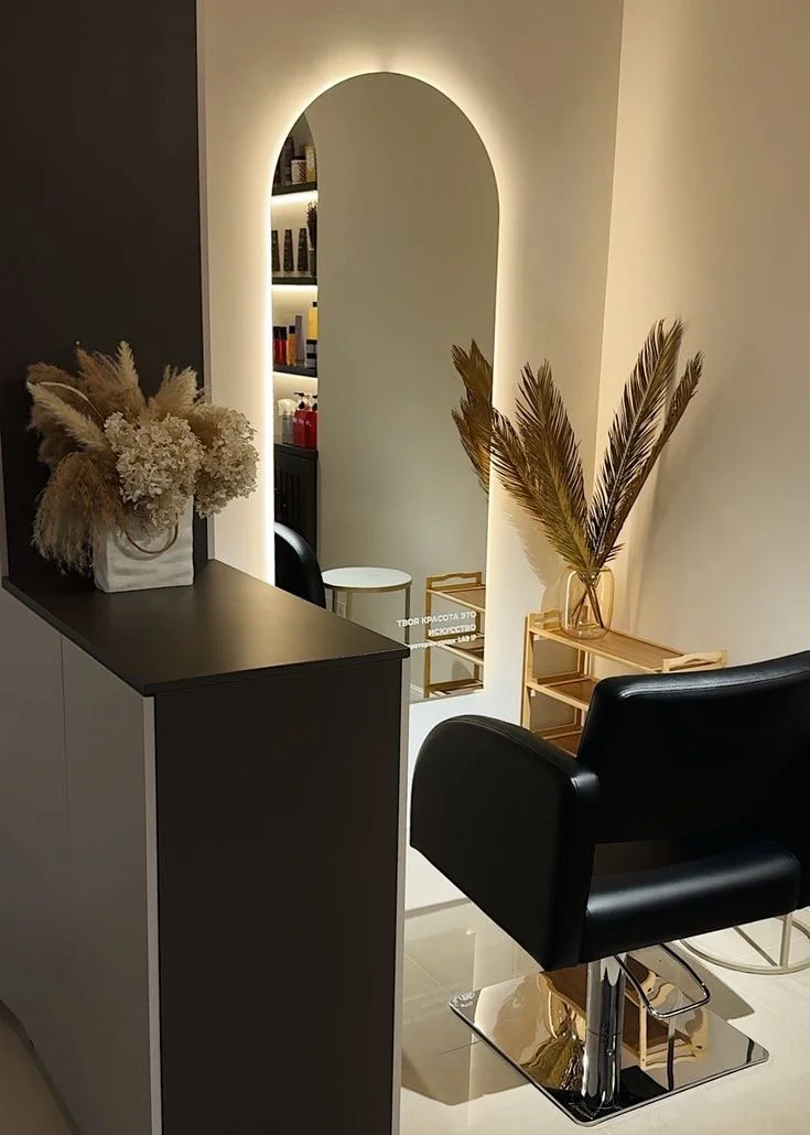 A salon or beauty studio with a black styling chair, a wooden shelf with dried palm leaves, a vase with pampas grass, a mirror with backlighting, and a glimpse of a shelf with products in the background.