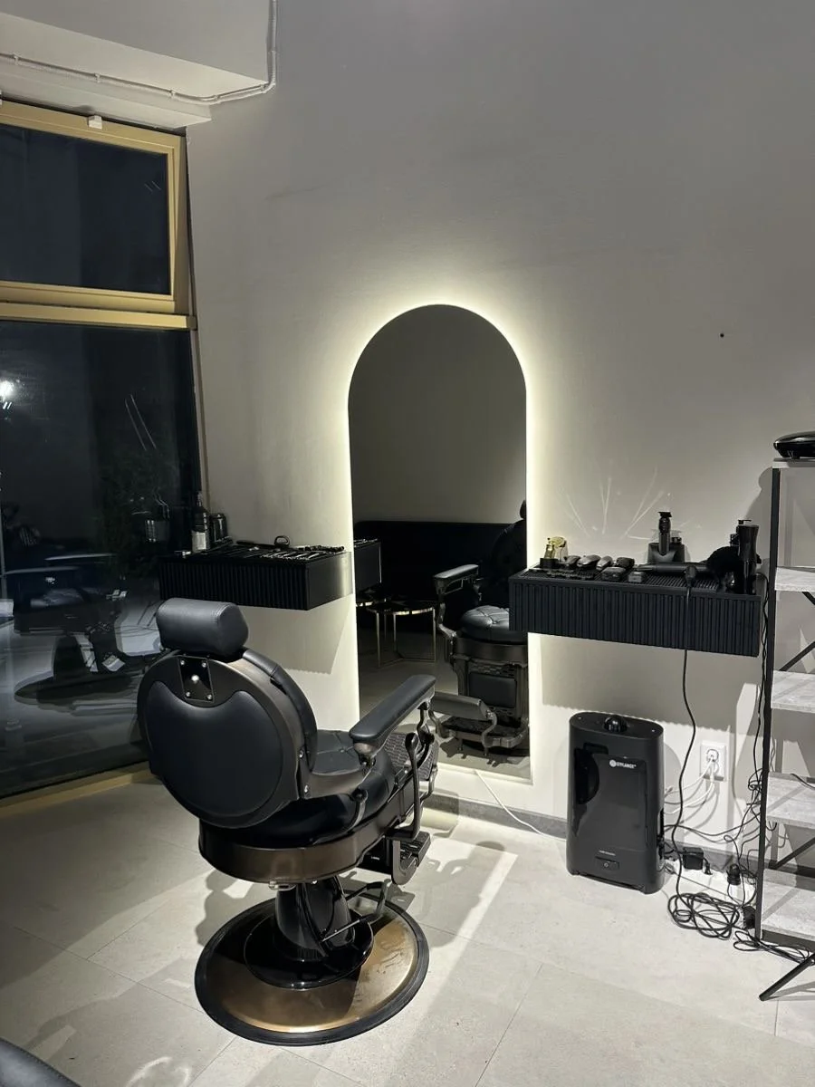 Barber shop with a barber chair facing a mirror, lighting around the mirror, and shelves with barber tools. Large window to the left with a reflection of the chair.