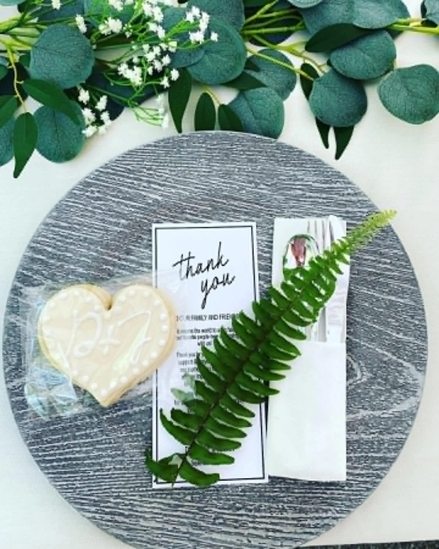 A round gray placemat on a white surface, decorated with green leaves and small white flowers at the top, a heart-shaped cookie with white icing and the word 'Thank' written on it, and a card with a picture and black text.