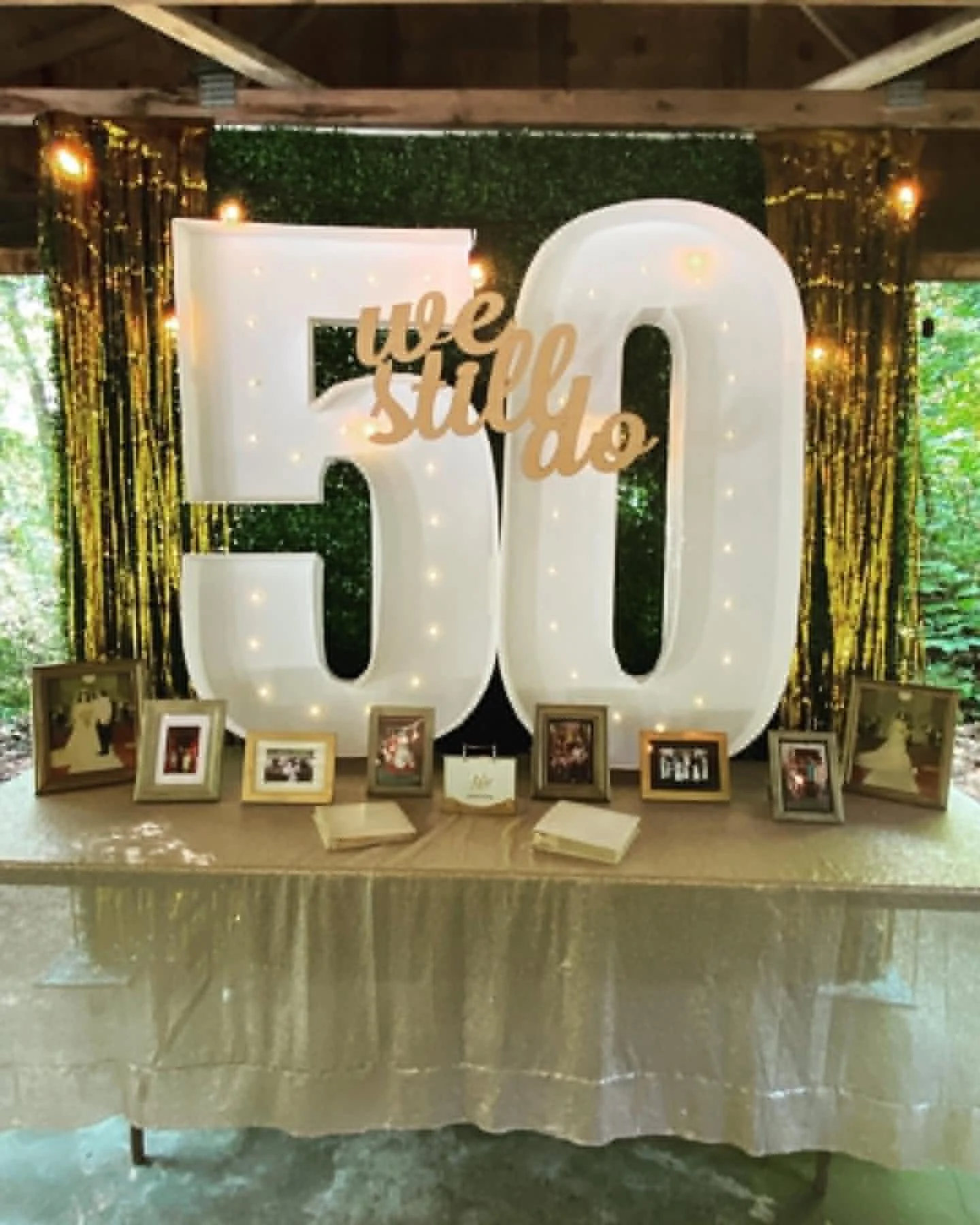 Wedding reception table with large illuminated '50' numbers, a sign reading 'we do still,' framed photographs, and a green leafy backdrop with string lights.