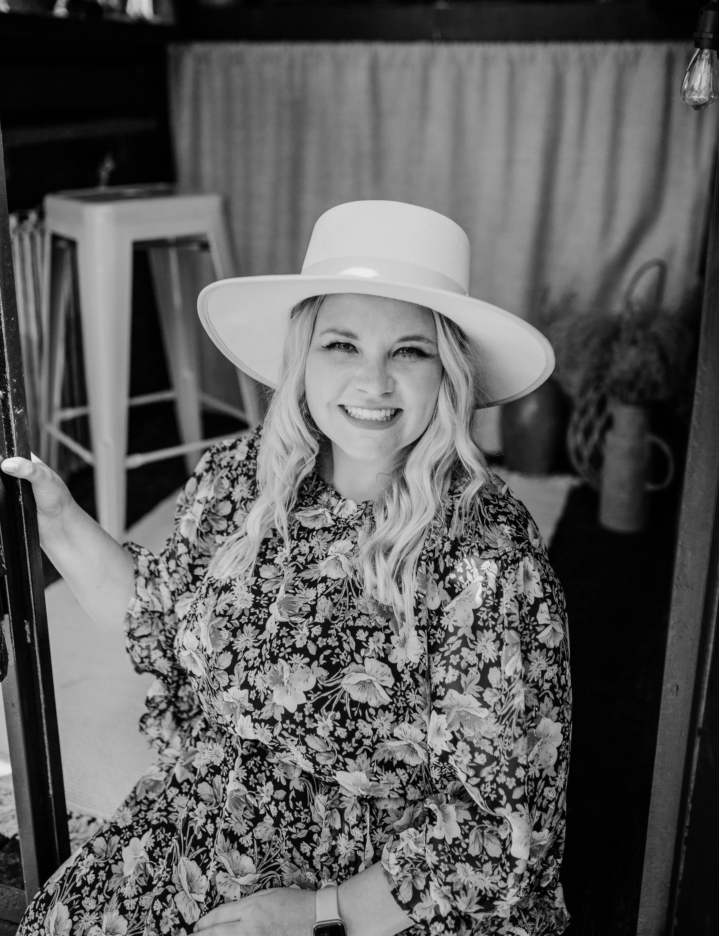 A woman with long, wavy blonde hair wearing a wide-brimmed hat and a floral dress, smiling and holding a window frame in an indoor setting.