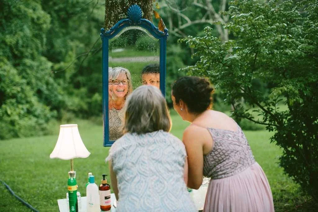 Two women applying makeup or styling hair in front of a mirror outdoors surrounded by greenery, with a white lamp and various bottles on a table nearby.