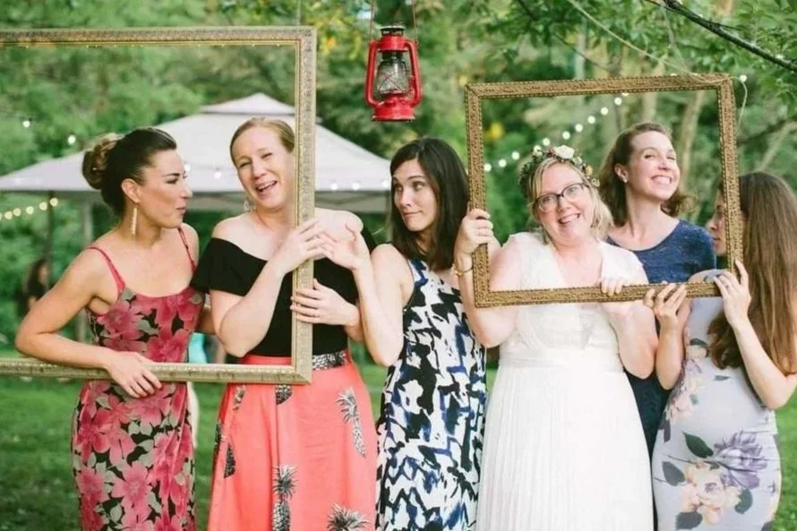 Group of women at an outdoor gathering holding empty picture frames and smiling, with a red lantern hanging above them.