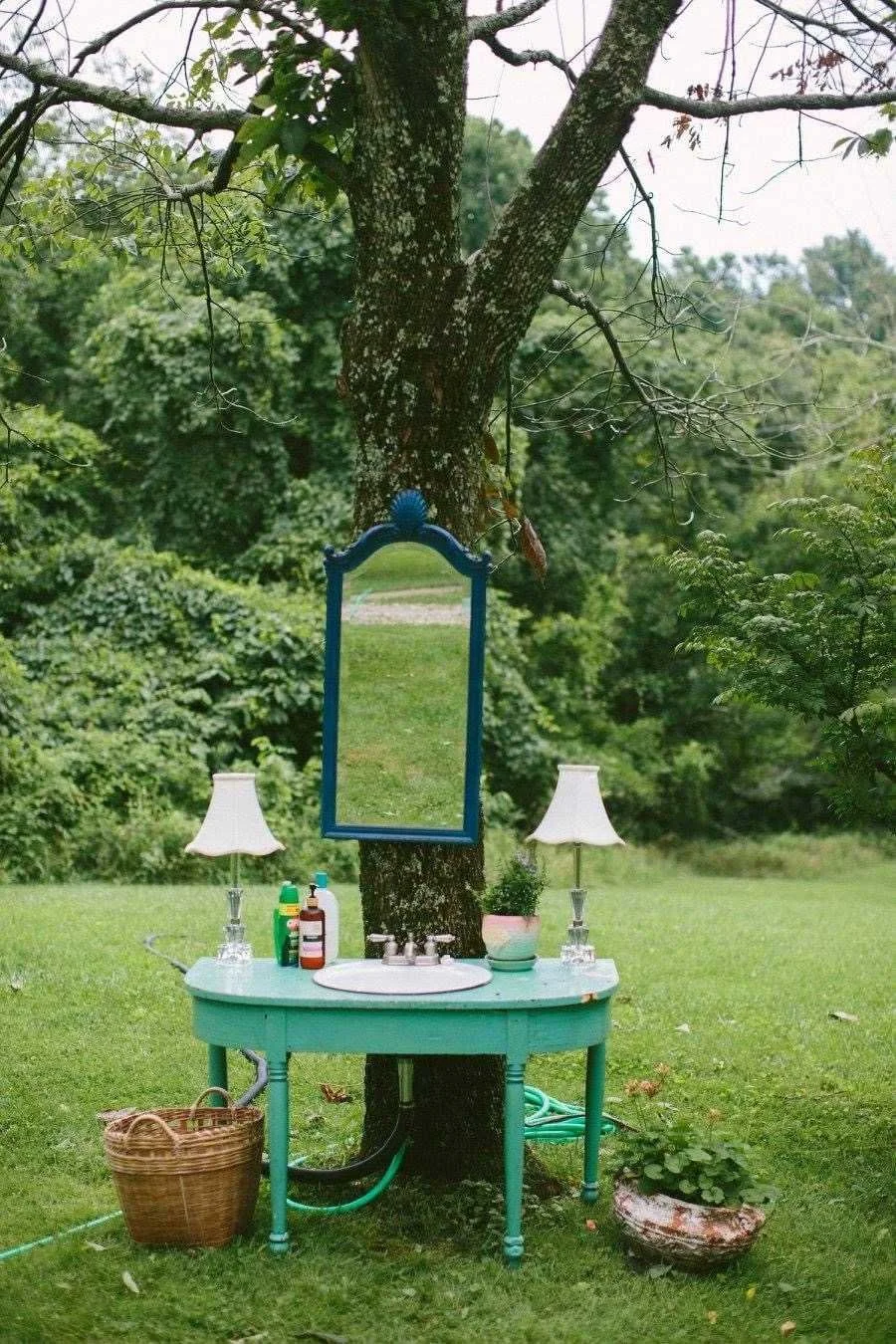 An outdoor scene with a tree, a turquoise sink, two white lamps, a mirror, and various bottles, in a grassy garden setting.