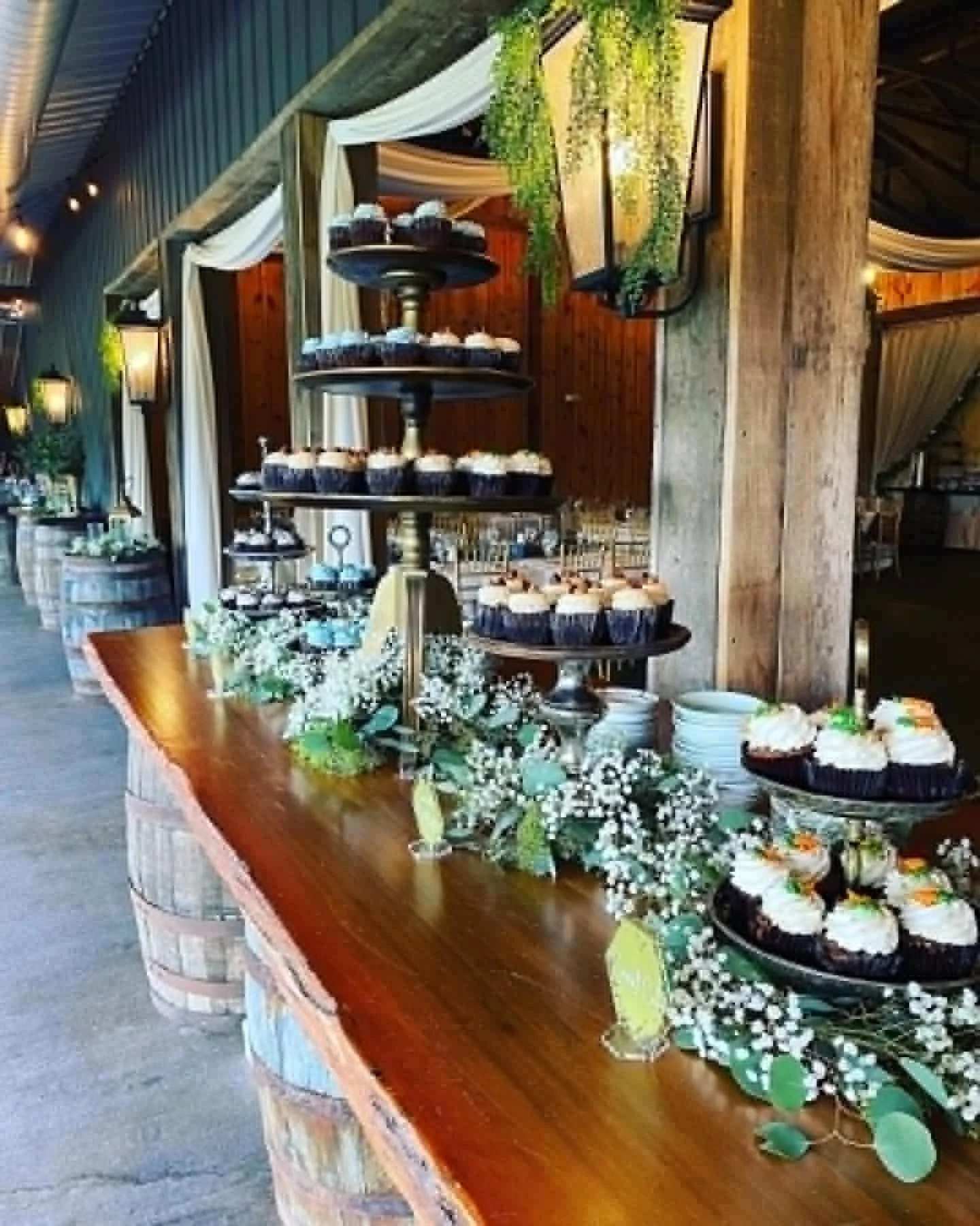 Table decorated with cupcakes, flowers, greenery, and a tiered stand with additional desserts at a rustic event space.