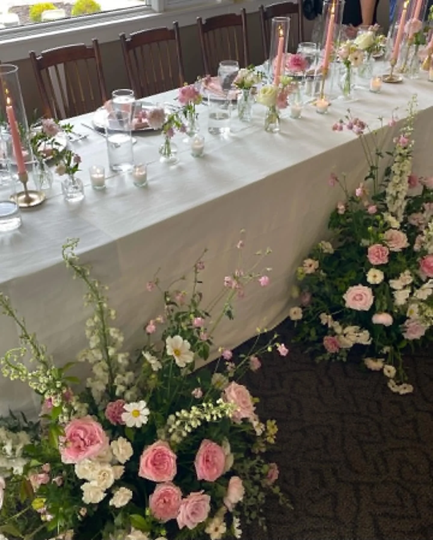 A long dining table decorated with pink candles, glassware, and small floral arrangements, set up in front of a window with flowers and chairs along one side.