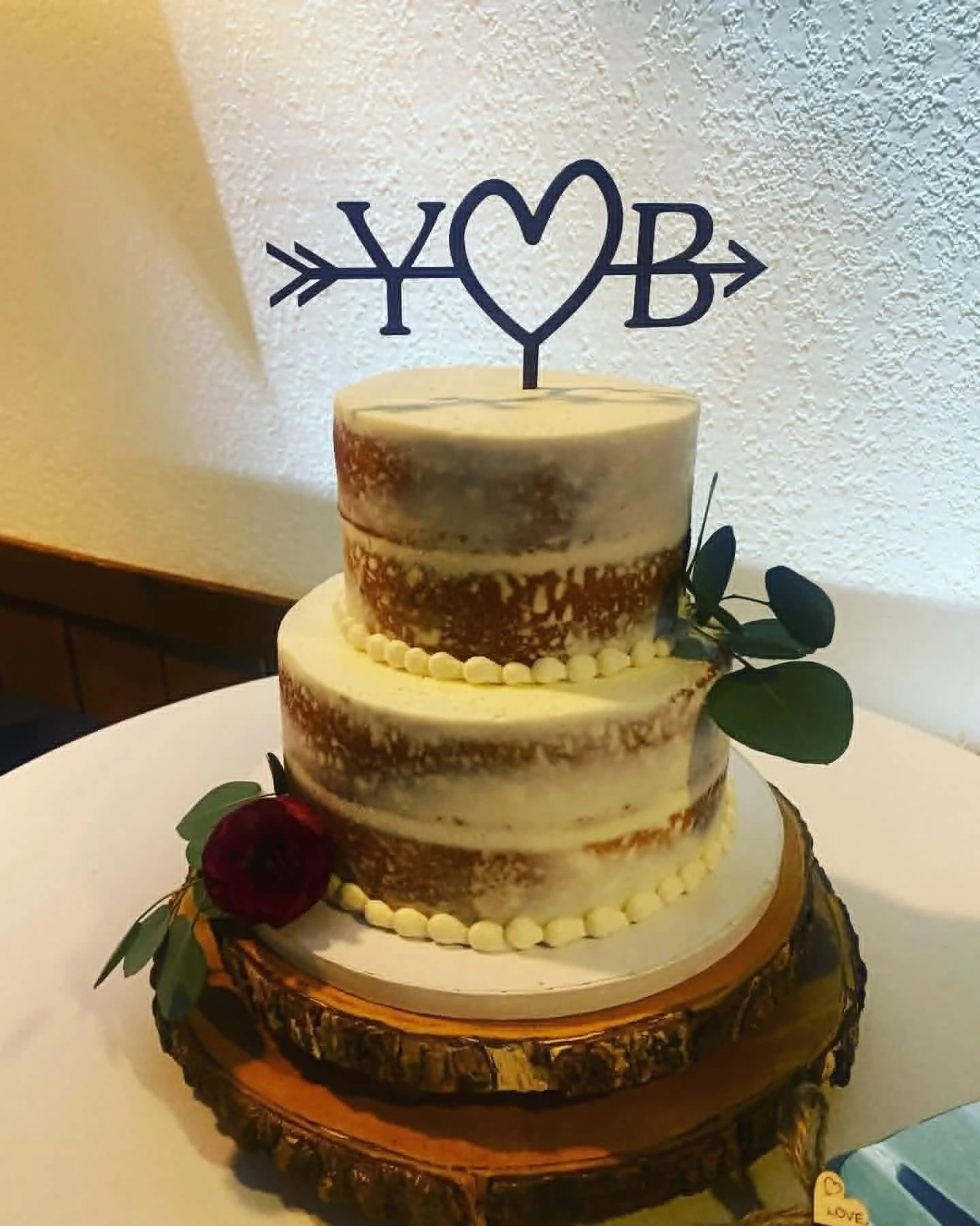 Three-tier wedding cake with a 'Love' cake topper, decorated with greenery and roses, on a wooden stand