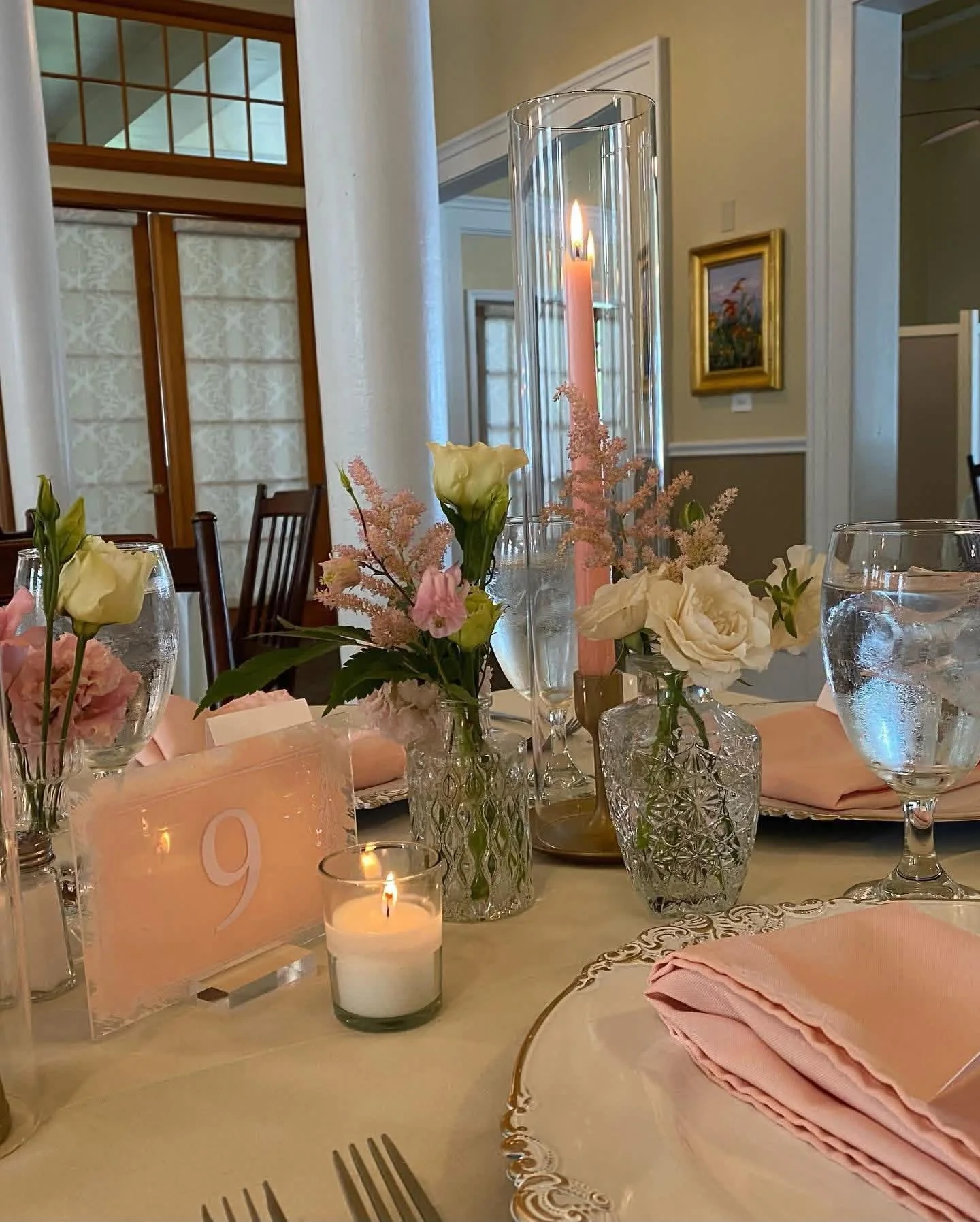 A formal dining table decorated with pink and white floral arrangements, pink candles, pink napkins, and a number 9 table card. There are water glasses and a lit candle in a small glass holder. The background shows a staircase and framed artwork on the wall.