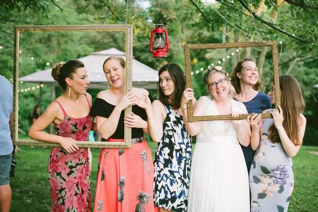 Group of women holding picture frames at an outdoor party, smiling and laughing, with string lights and trees in the background.