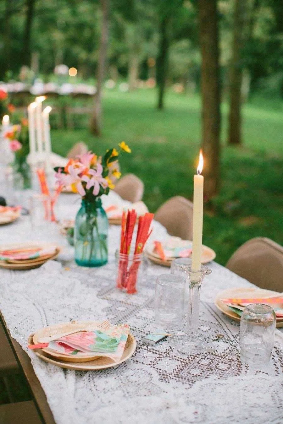 Outdoor dining table set with floral centerpieces, candles, and colorful napkins, in a lush green garden or park.