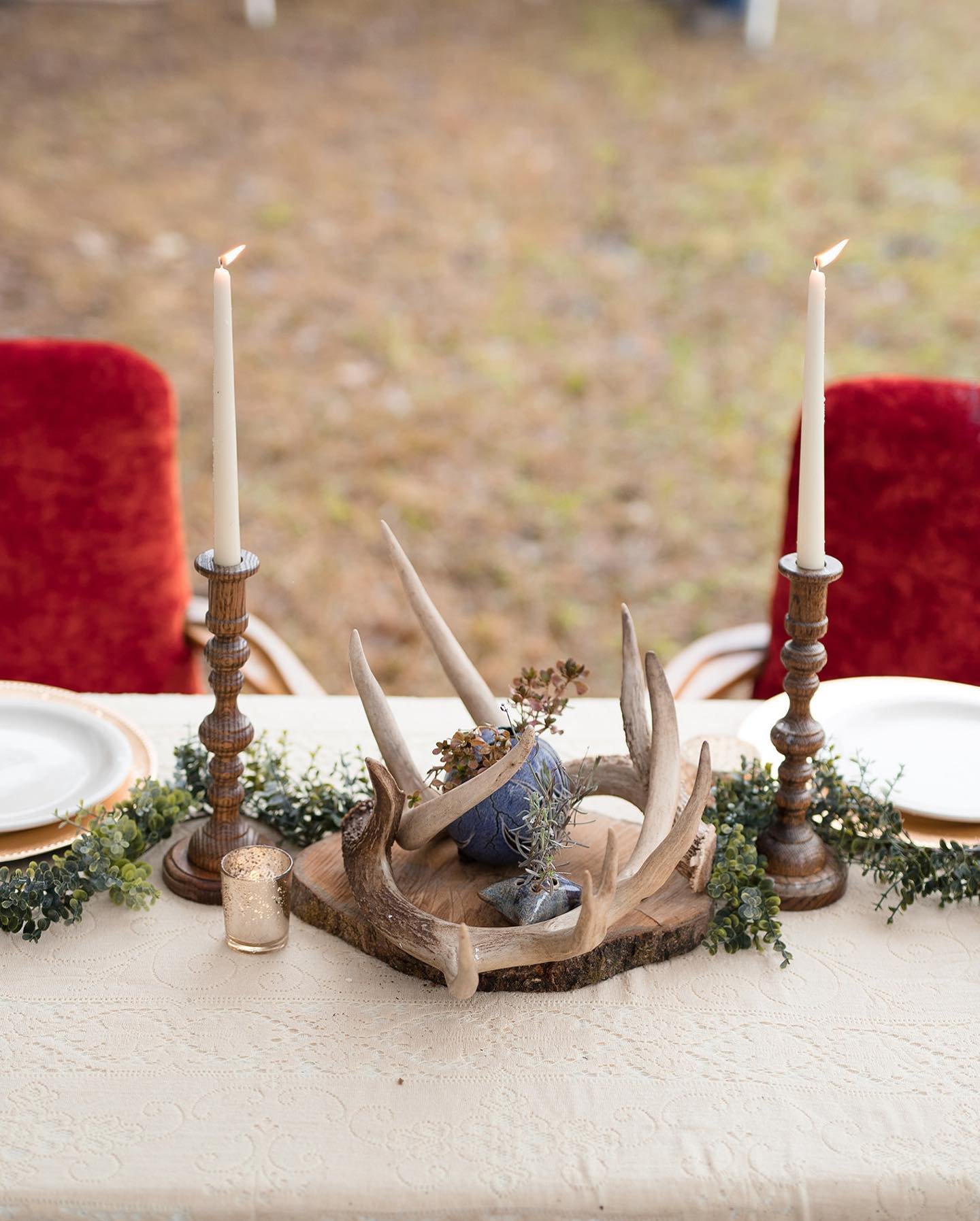 A rustic table setting with candles, greenery, antlers, and decorative stones, set outdoors with red cushioned chairs in the background.
