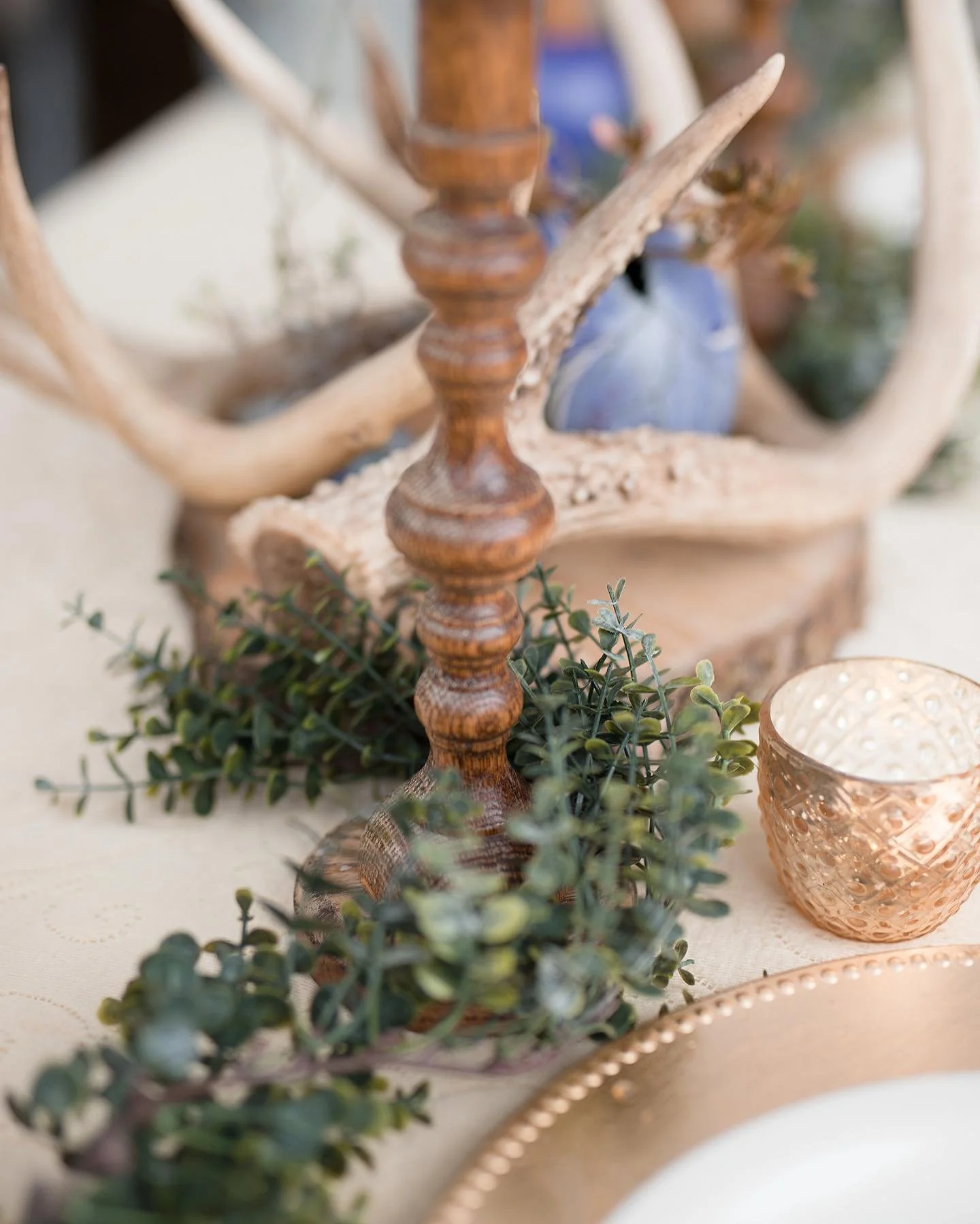 Decorative arrangement with a wooden candlestick, green foliage, a blue vase, a peach-colored candle holder, and a driftwood piece behind.