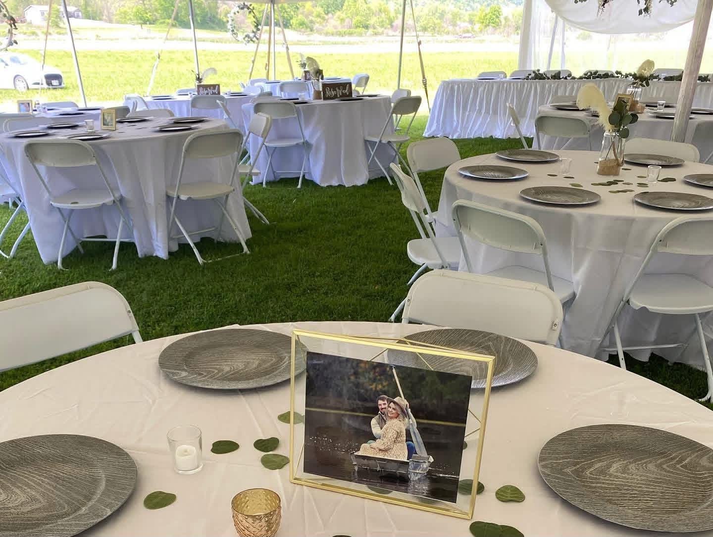 Wedding reception tent with round tables decorated with white tablecloths, wooden plates, small candles, and a framed photograph of a couple on a boat with an oar, greenery, and open outdoor landscape in the background.