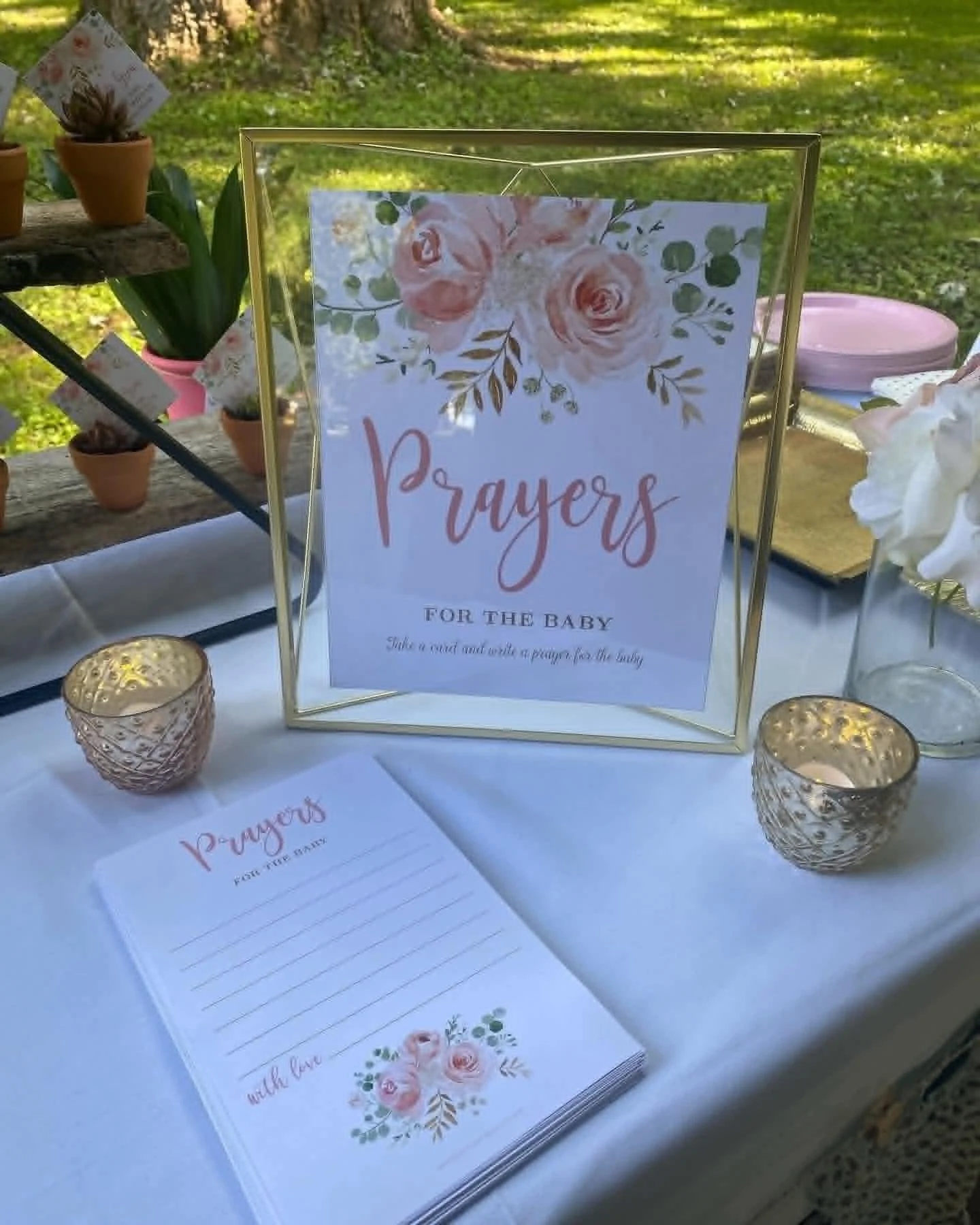 A sign with the word "Prayers" and a floral design, set on a table with small candles and prayer cards, outdoors with green grass and trees in the background.