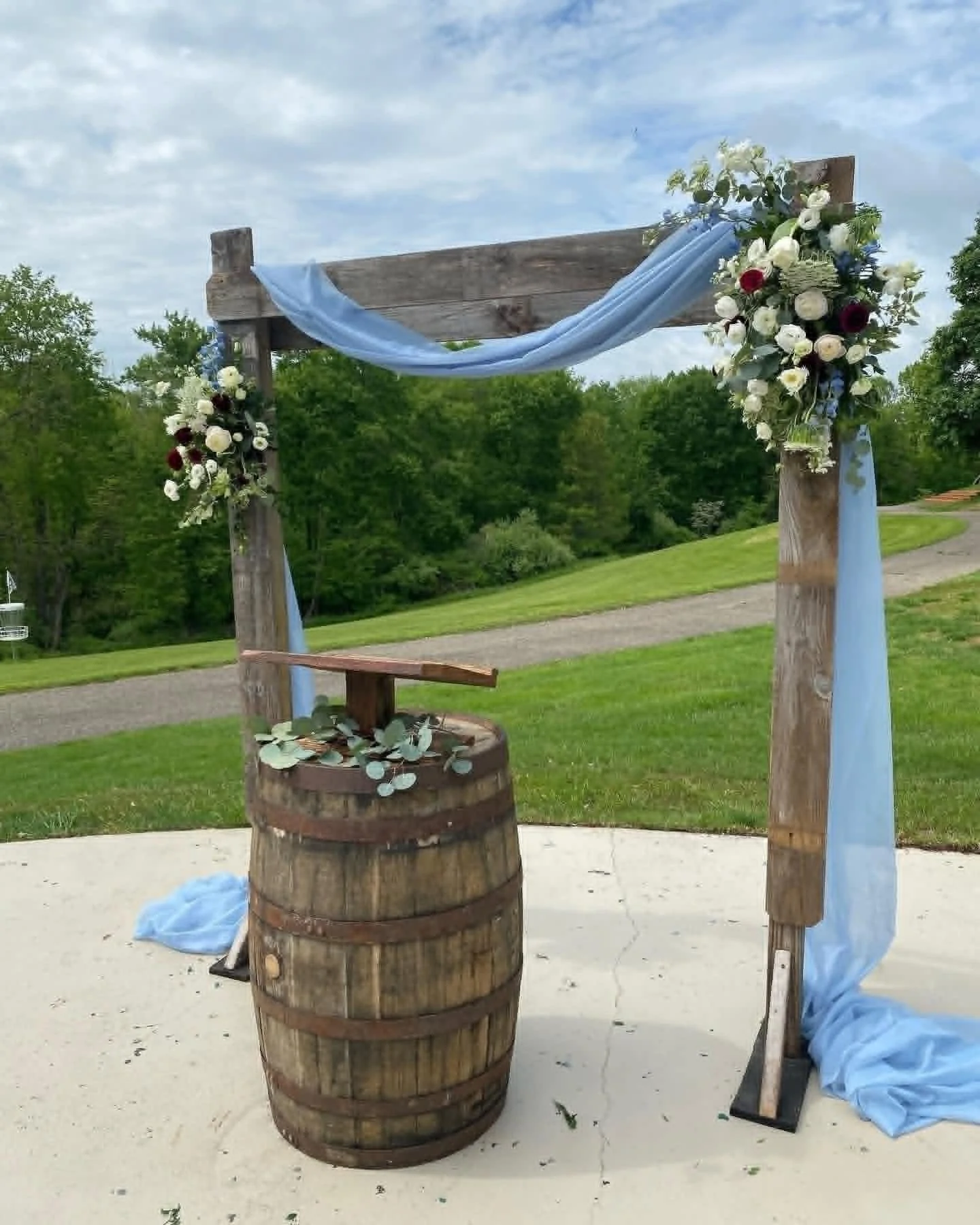 Wedding arch decorated with blue fabric and white and dark red flowers, empty and set outdoors on grassy area.