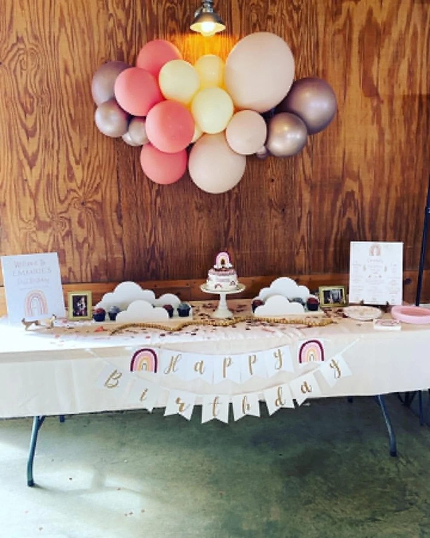Decorated dessert table with pastel balloons arranged on a wooden backdrop, colorful cupcakes, a rainbow cake, and a 'Happy Birthday' banner featuring rainbows and clouds.