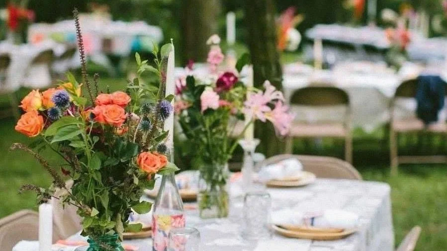 A table decorated with colorful flower arrangements in vases and set with plates, napkins, and glasses outdoors under a green canopy.