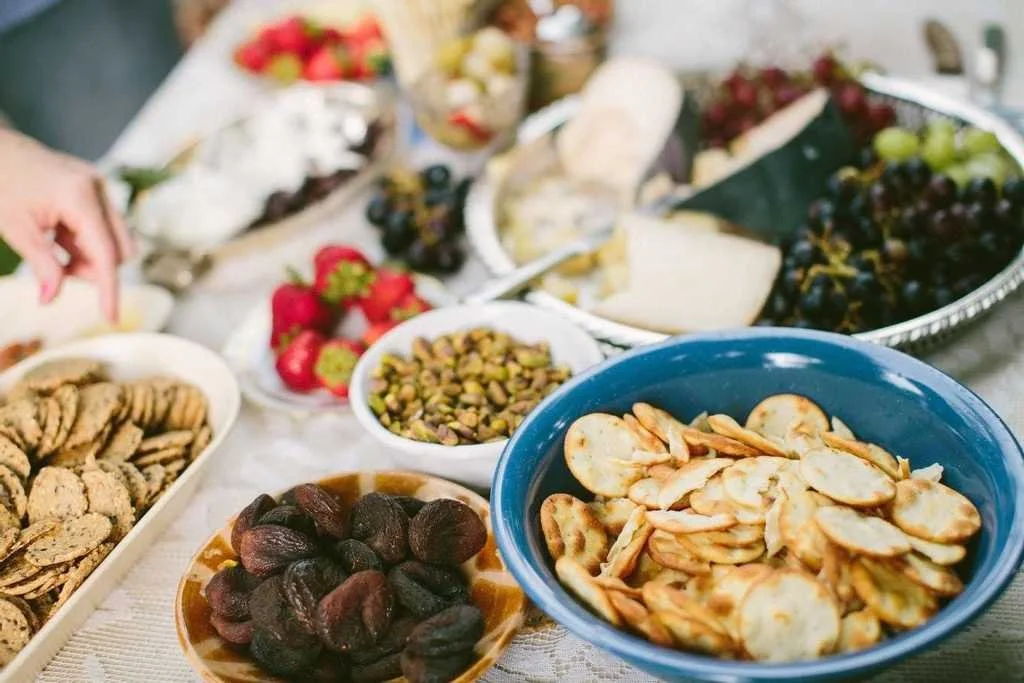 An assortment of snacks and fruits on a table, including cookies, dried apricots, pistachios, strawberries, grapes, and crackers.
