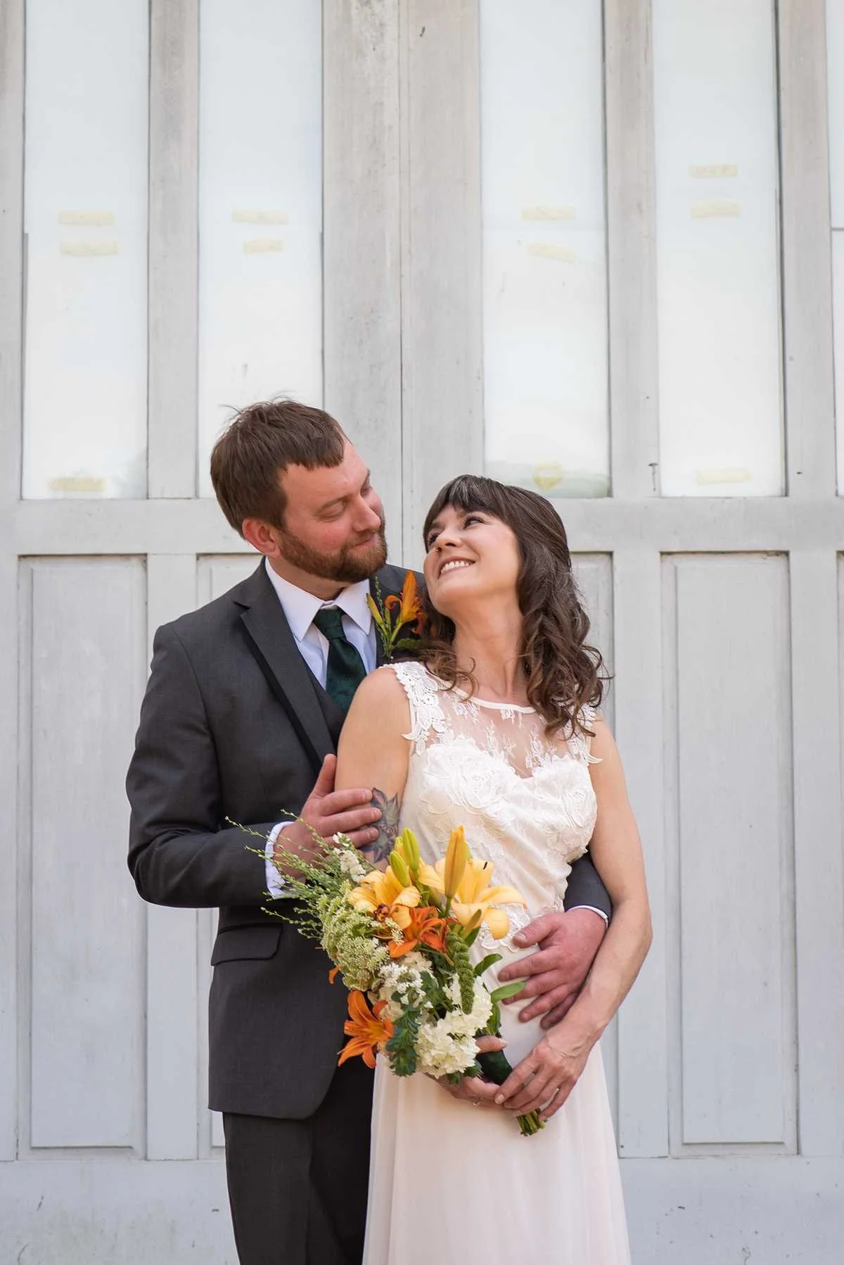 A newlywed couple embraces in front of a white wooden wall. The groom, dressed in a dark grey suit, holds the bride gently by her waist, while the bride, in a white lace wedding gown, holds a colorful bouquet of orange, yellow, and white flowers. Both are smiling and gazing affectionately at each other.
