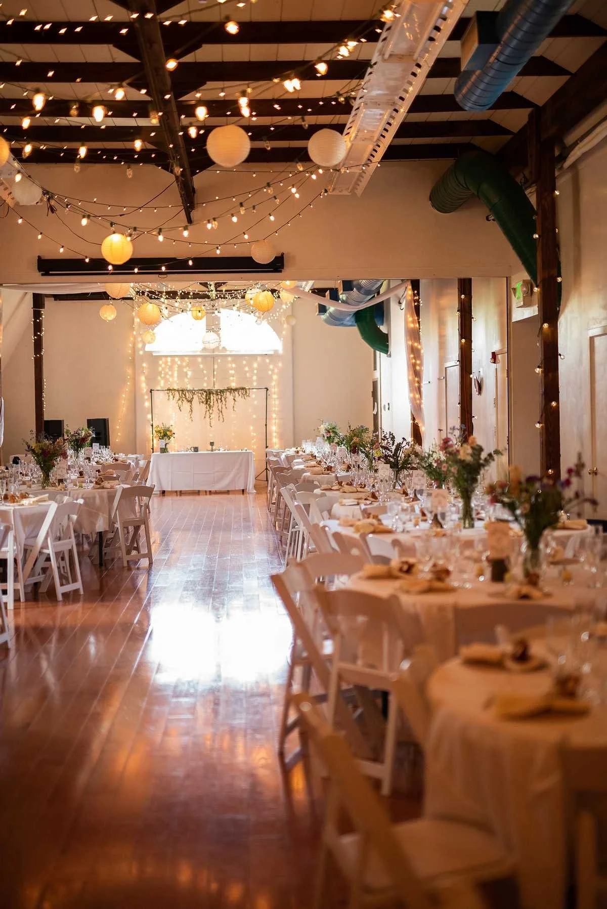 A decorated banquet hall set for a formal event with long tables, white chairs, floral centerpieces, string lights, and paper lanterns hanging from the ceiling, with a backdrop featuring fairy lights and greenery.