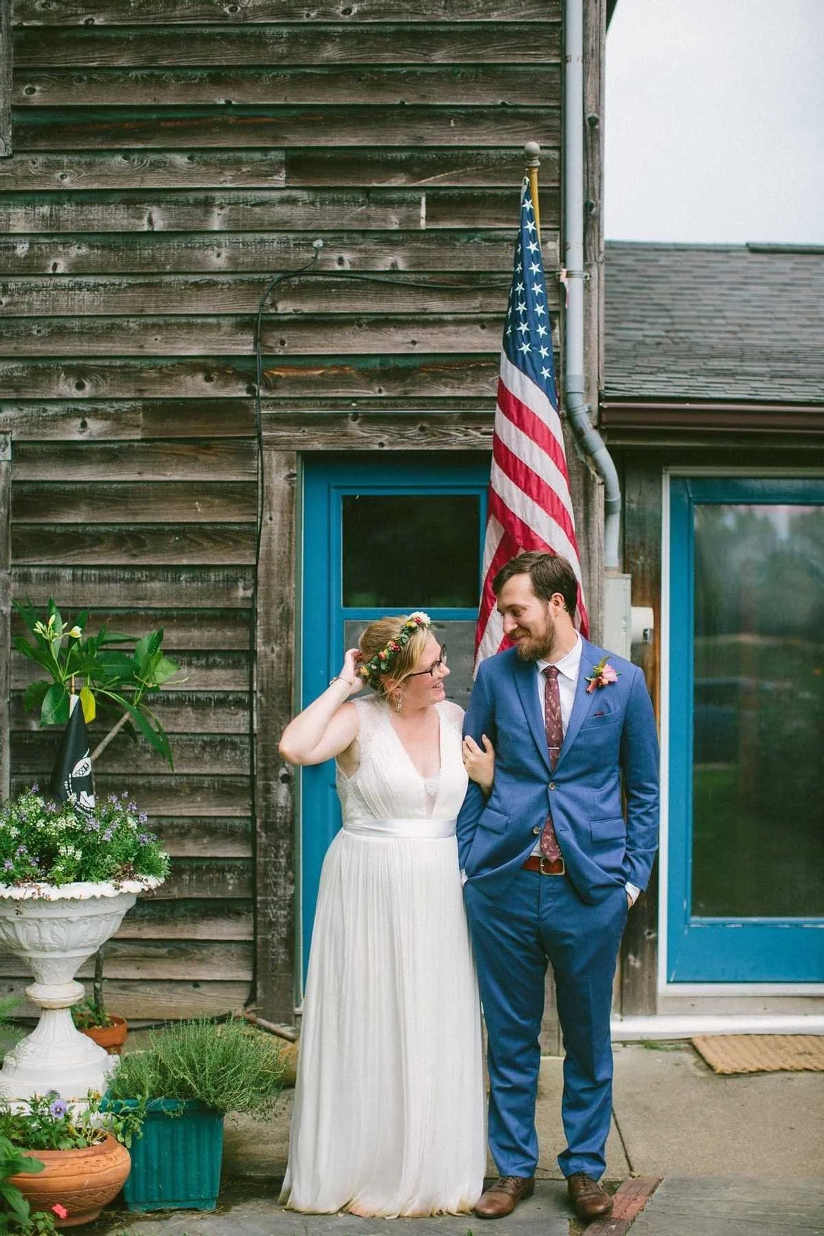 A bride and groom standing outside a rustic wooden building. The bride wears a white dress and floral crown, and the groom wears a blue suit. They are smiling and looking at each other. An American flag hangs behind them, and there are potted plants and flowers nearby.