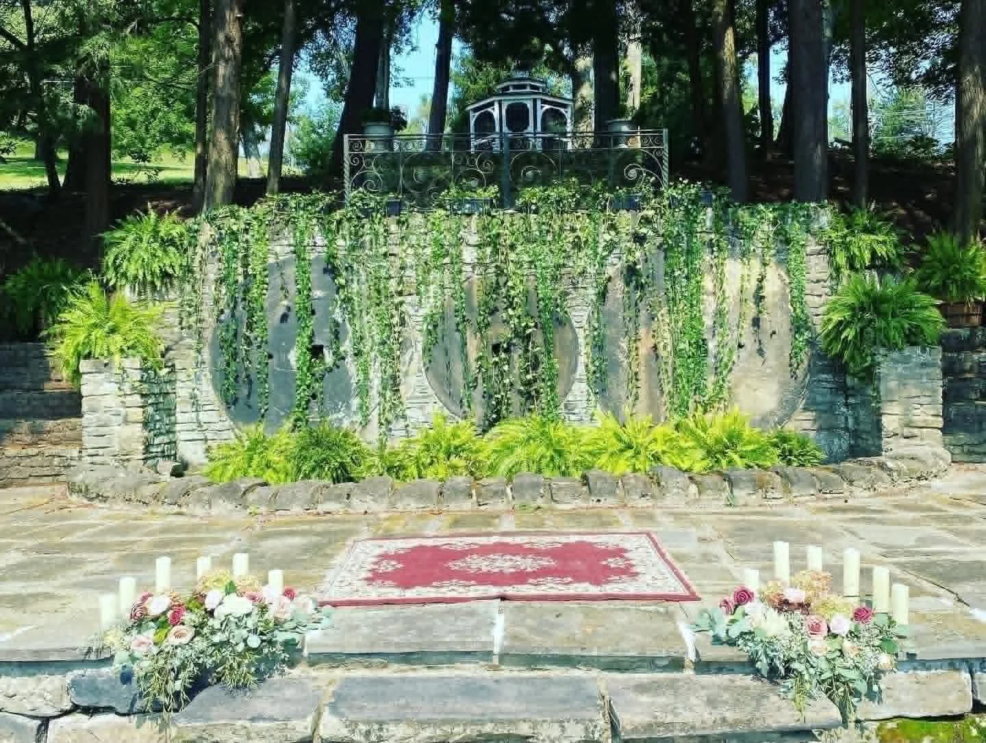 Outdoor wedding altar decorated with flowers and candles, with a stone wall and lush greenery in the background.