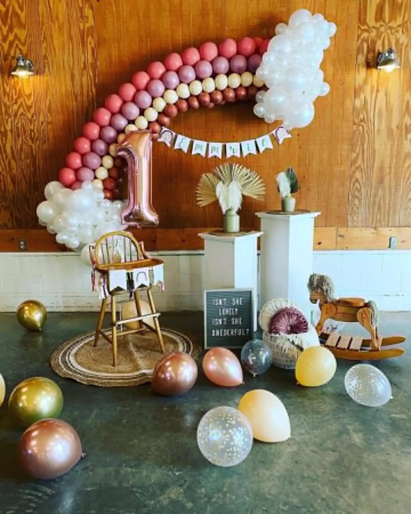 Birthday party decorations with a large pink and gold balloon number 1, a rainbow-shaped balloon arch, and various balloons scattered on the floor. There are small white pedestal tables with festive decorations and a wooden wall in the background.