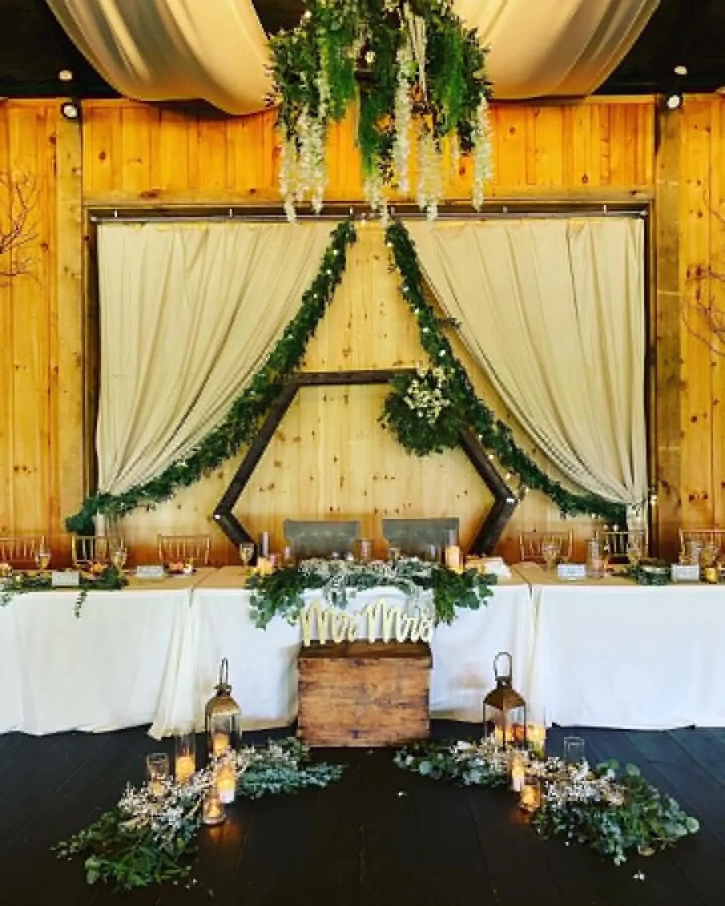 Wedding reception table decorated with white tablecloths, green foliage, and white flowers, with a backdrop of cream curtains and wooden walls, including a large star frame and floral hanging centerpiece.