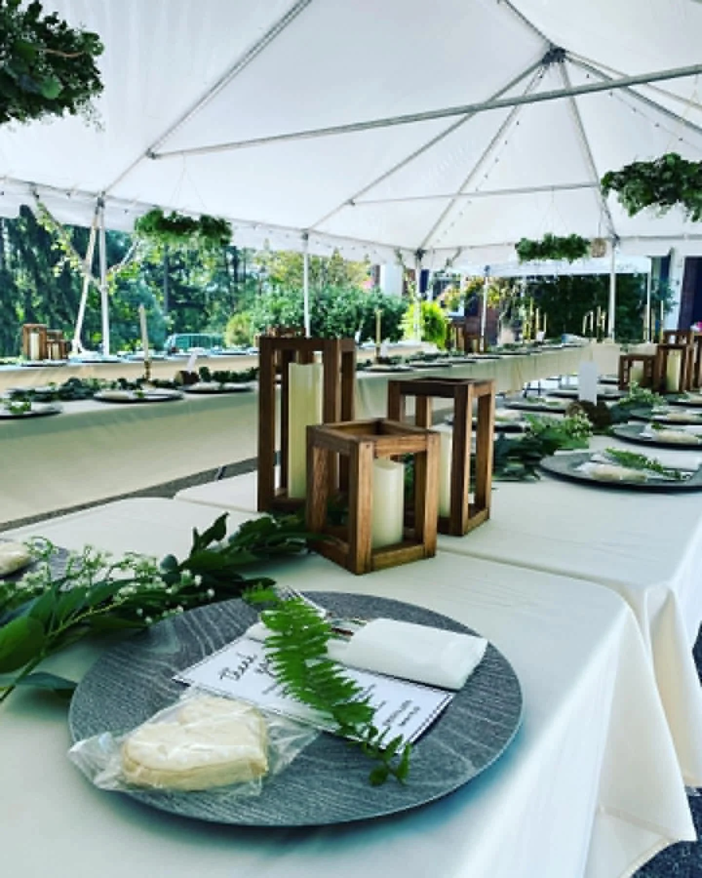 Wedding reception table setup with white tablecloths, green foliage decor, and a menu card. A plate of cheese and crackers, with a fork, napkin, and greenery, is in the foreground. The scene is outdoors under a large white tent with trees and greenery outside.