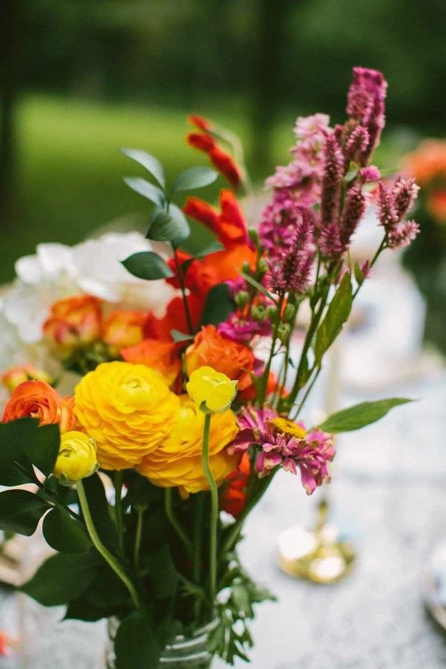 A colorful bouquet of various flowers including yellow, orange, pink, and purple blossoms with green leaves, arranged in a glass vase on a table outdoors.
