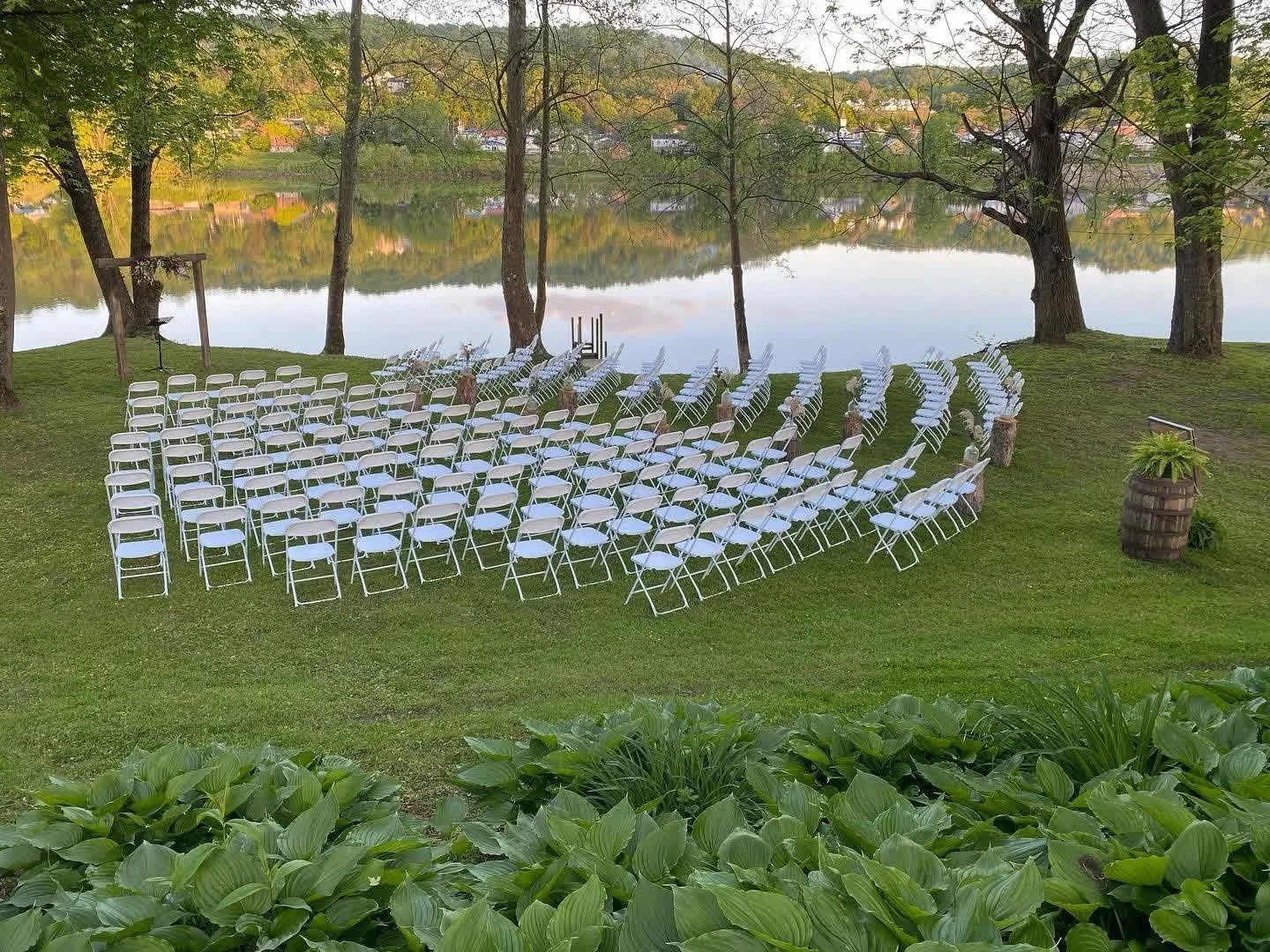 Outdoor wedding setup with rows of white chairs facing a lake, surrounded by trees and greenery, with scenic water and hills in the background.