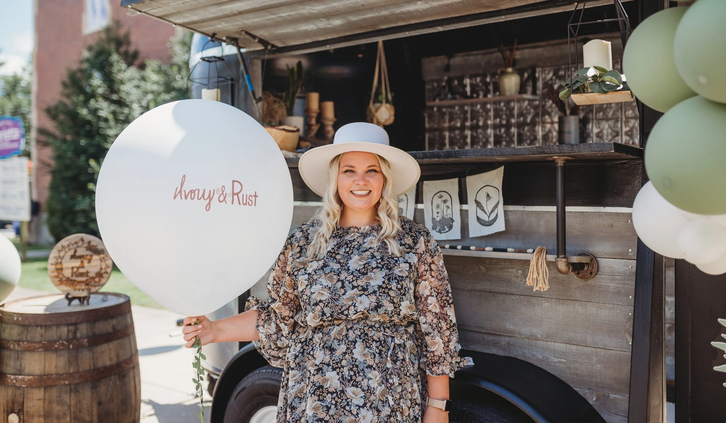 A smiling woman holding a white balloon with the words "Ivory & Rust" written on it, standing in front of a vintage food truck decorated with ivy and candles, outdoors on a sunny day.