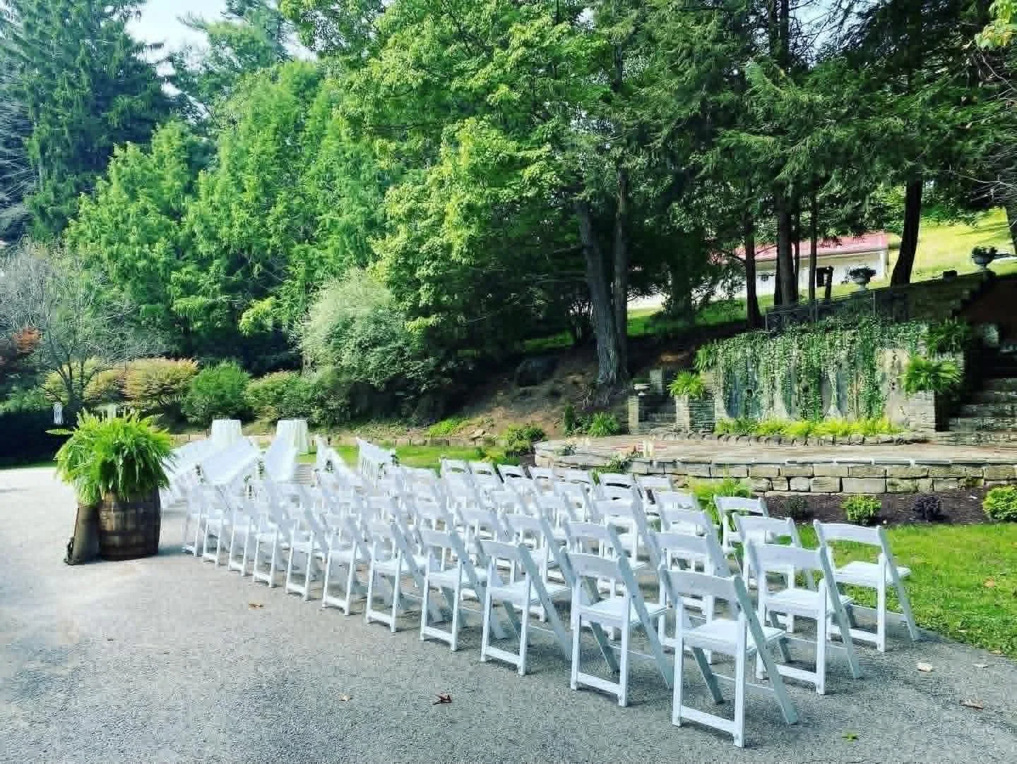 Outdoor wedding setup with rows of white chairs on a paved surface, potted plants, and lush green trees and shrubs in the background.