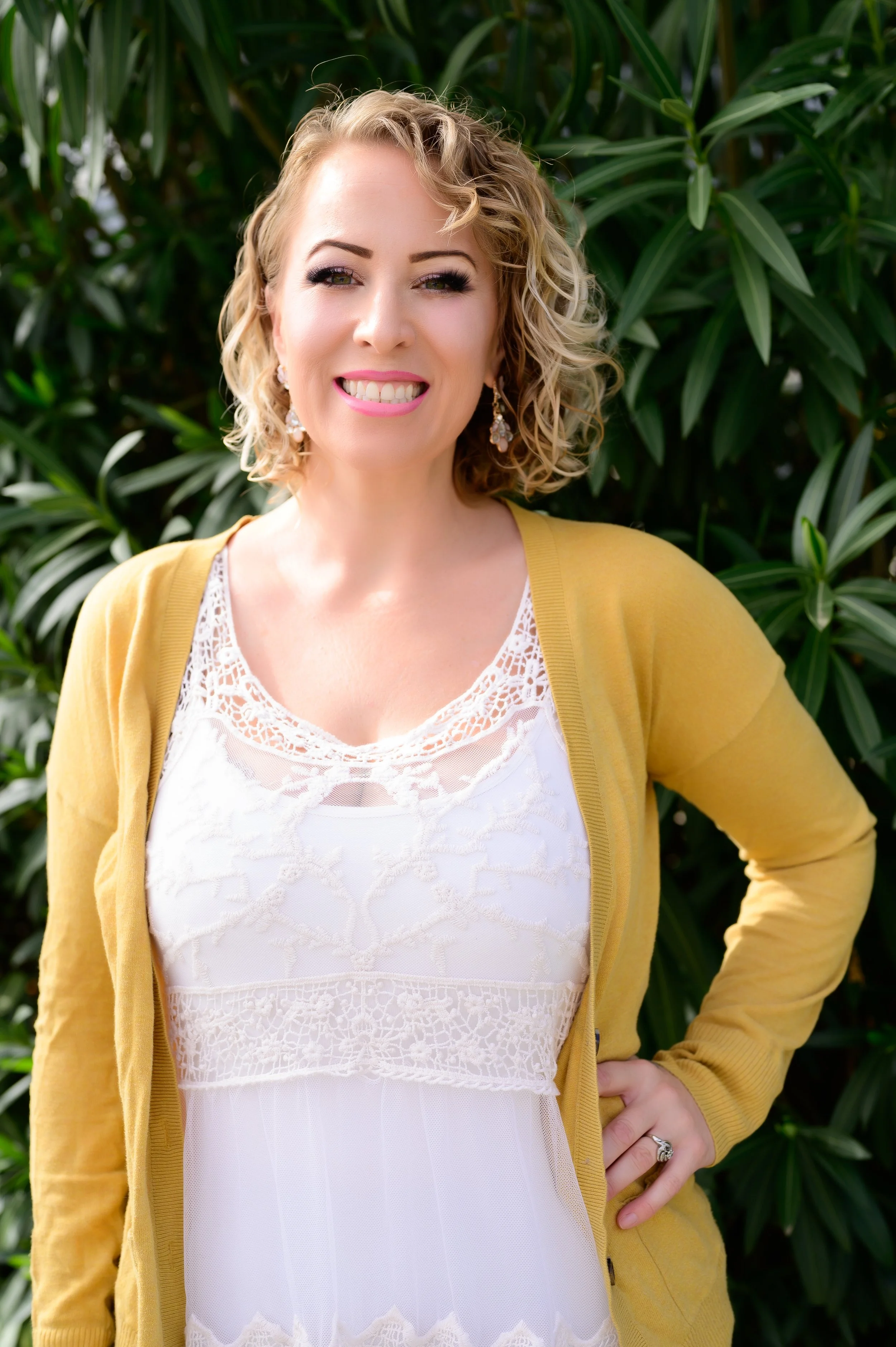A smiling woman with curly blonde hair, wearing earrings, a white lace top, and a yellow cardigan, stands outdoors in front of green foliage.