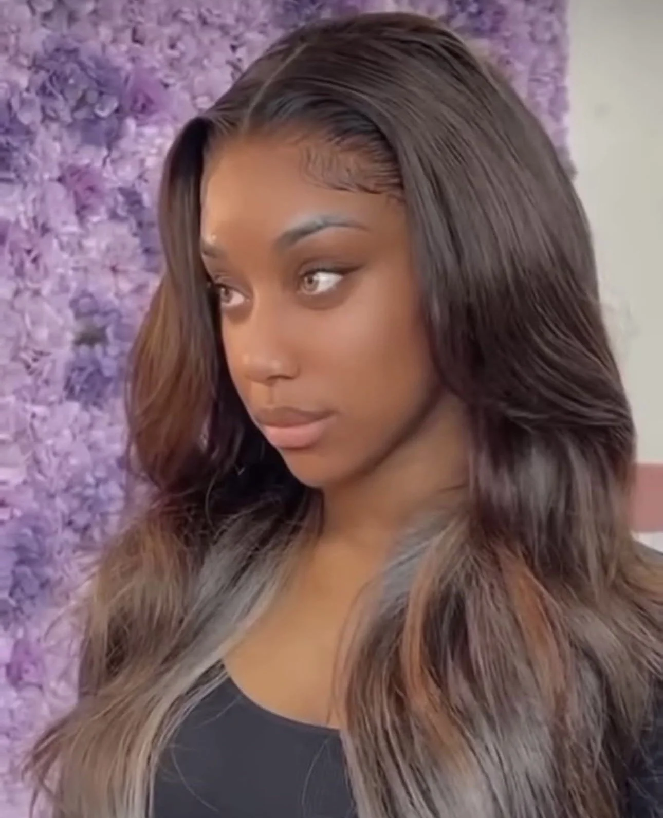 A woman with long, wavy hair that is ombre from dark brown to caramel stands in front of a purple floral background.