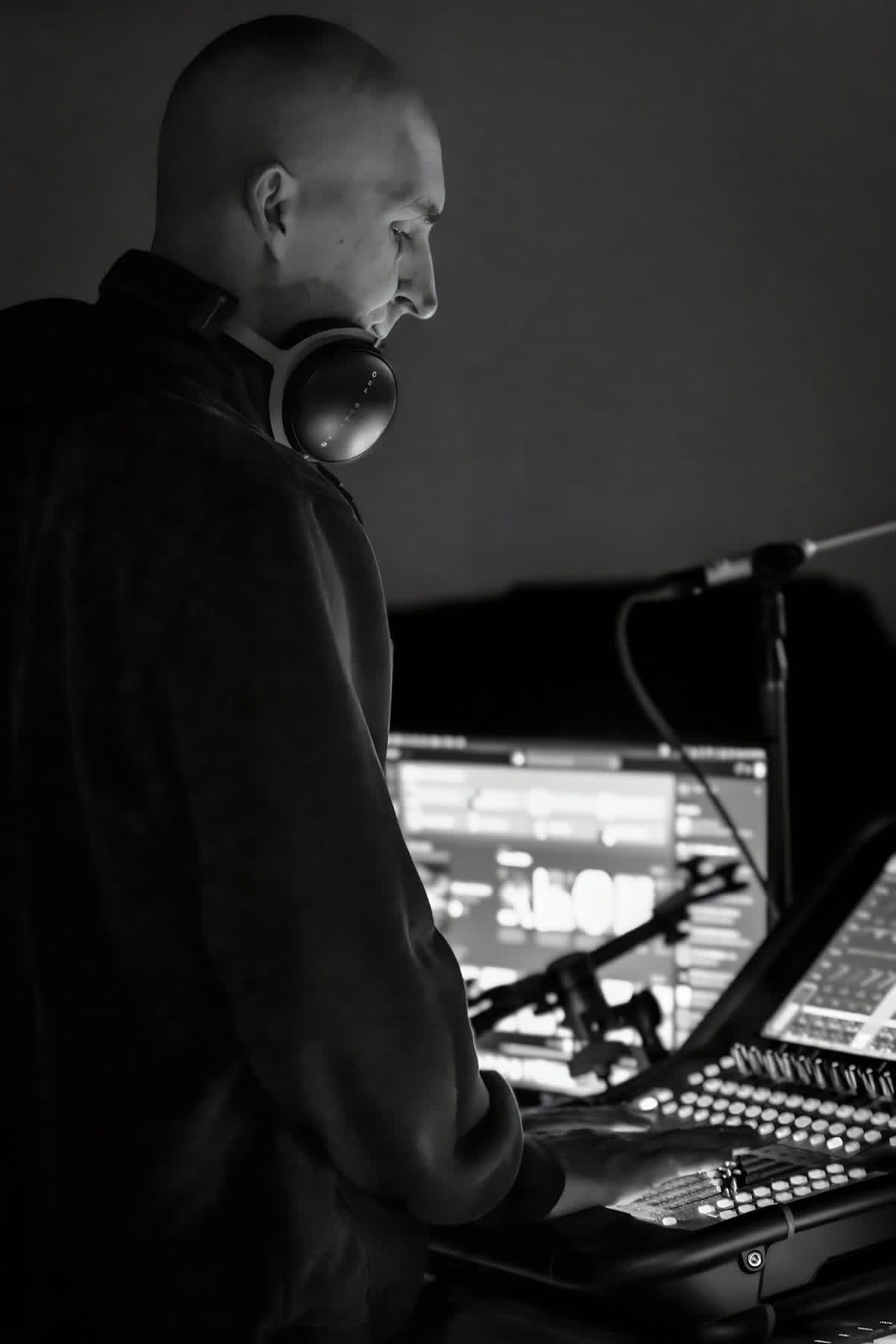 A person with a shaved head and headphones around their neck works at a mixer or soundboard with multiple screens and controls in a dark room.