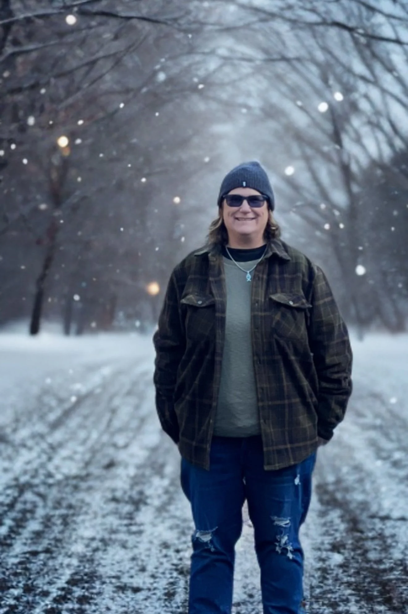 A smiling person wearing sunglasses, a gray beanie, a brown plaid jacket, and ripped jeans walking in a snowy, wooded area with bare trees and falling snow.