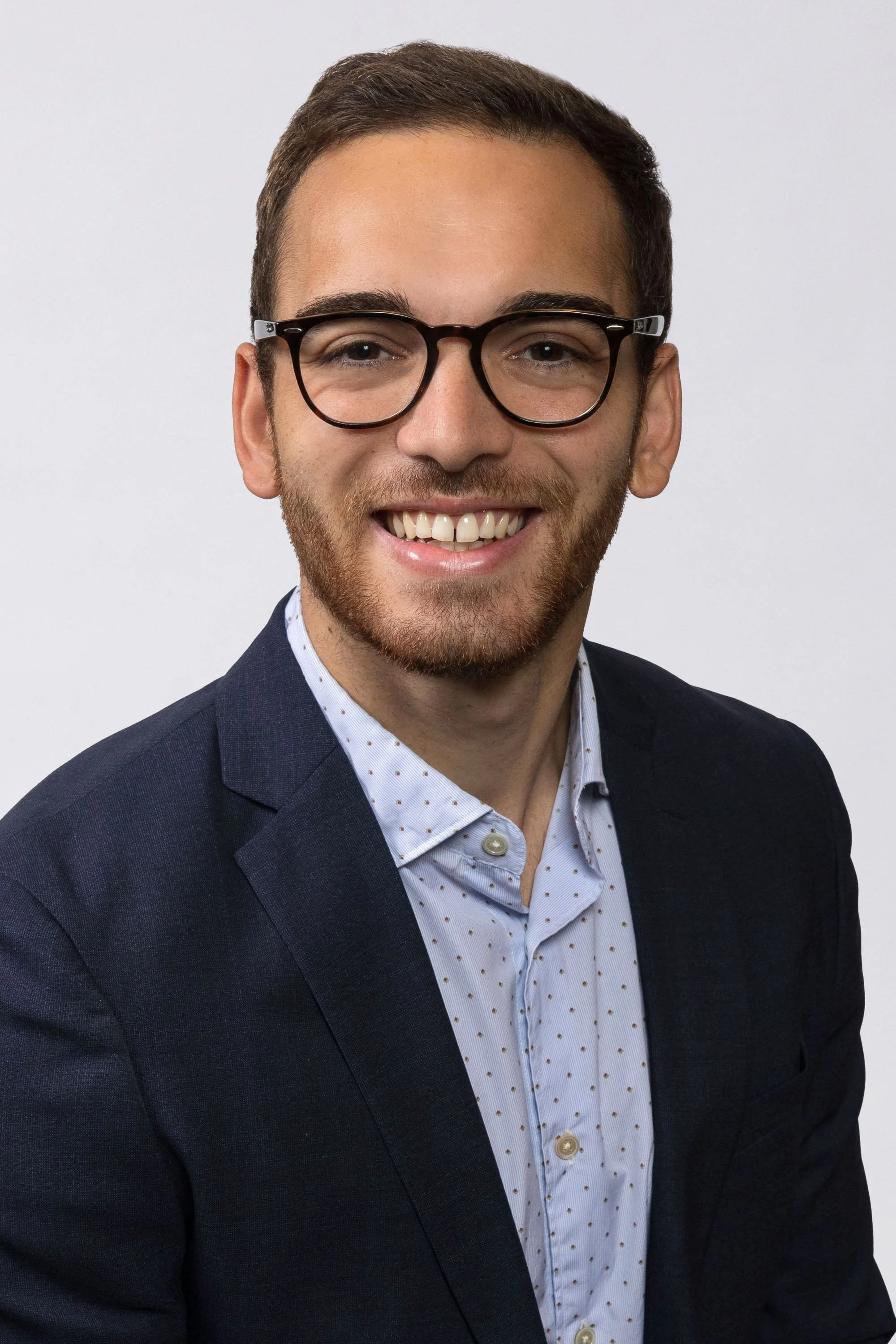 Headshot of a man with glasses, a beard, wearing a navy blazer and a light blue shirt with small darker dots, smiling against a plain white background.