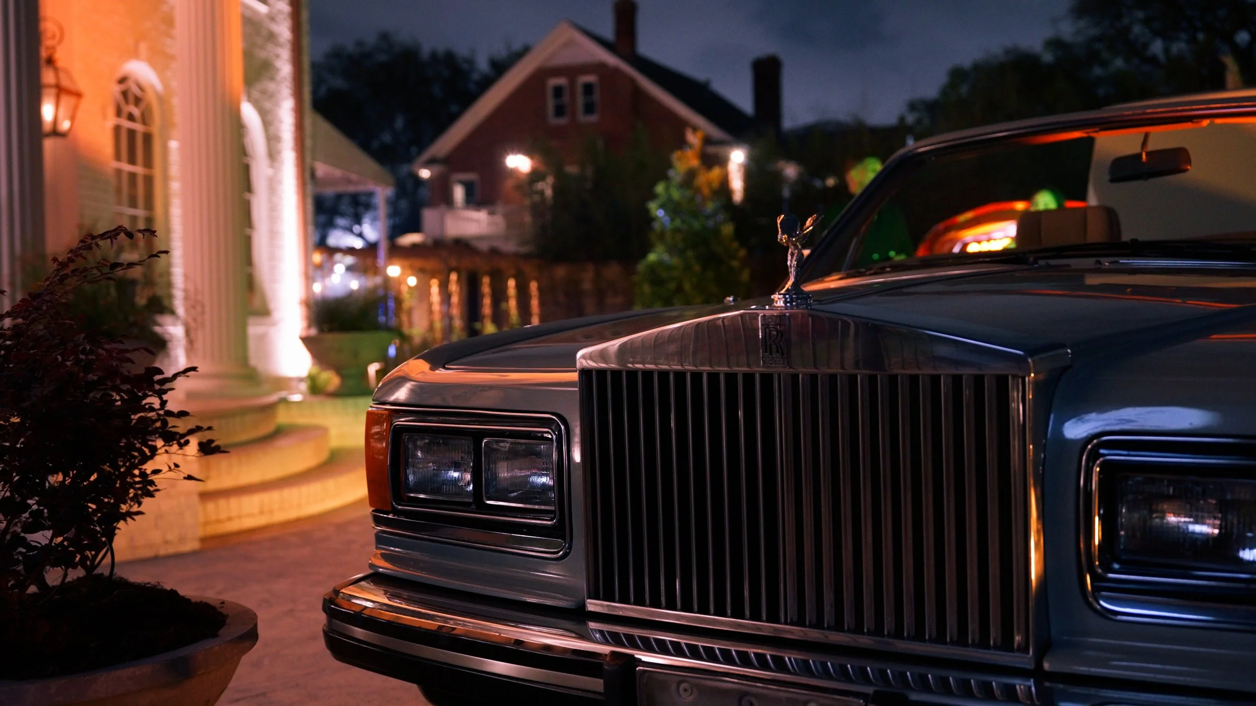 Close-up of the front of a vintage black Rolls-Royce car parked outside illuminated house at night.