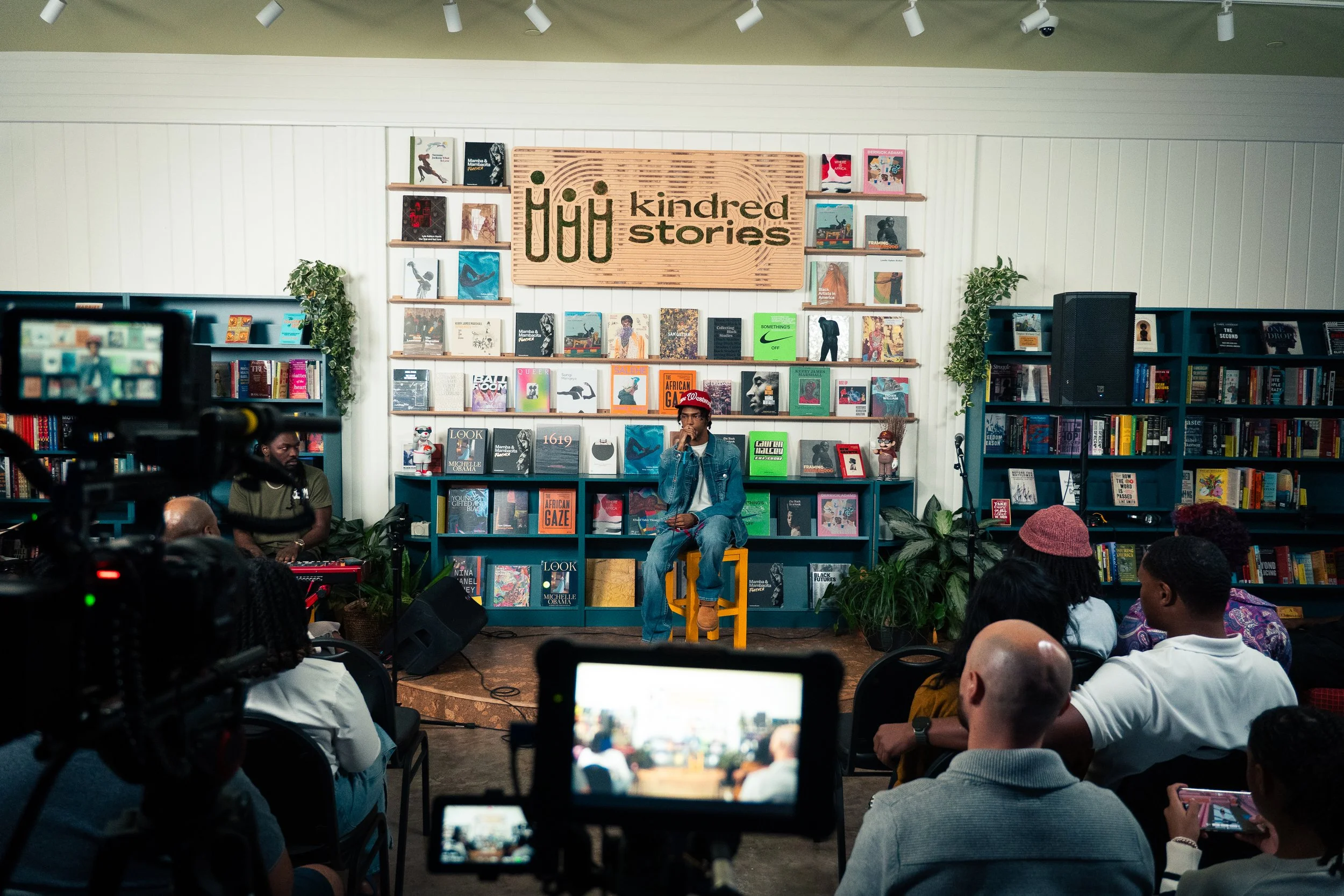 A person is seated on a yellow chair on a stage, speaking or singing into a microphone during an event at a bookstore called 'Kindred Stories' with an audience seated in front. The background features shelves filled with books and hanging plants.