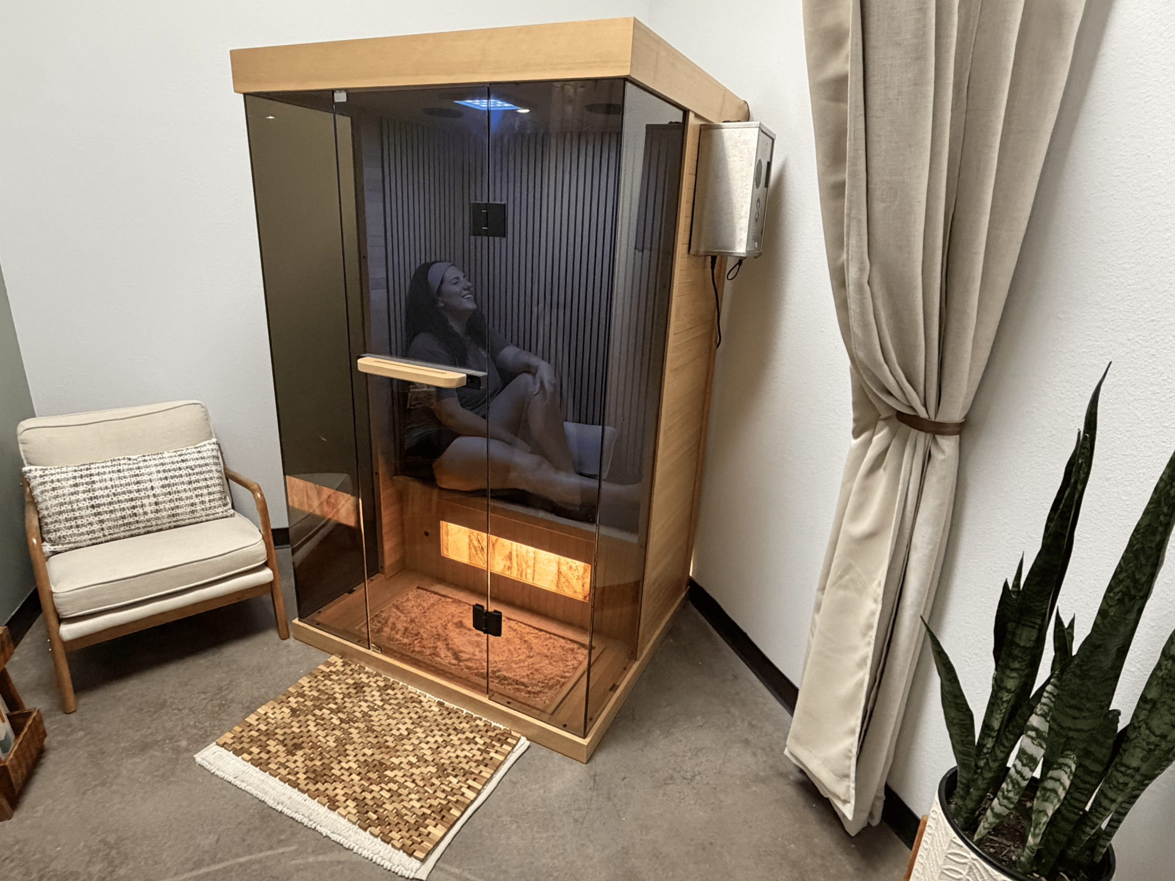 A woman sitting inside a halotherapy booth with glass walls in a spa. The halotherapy booth is made of wood inside. The woman is smiling and wearing a headband.