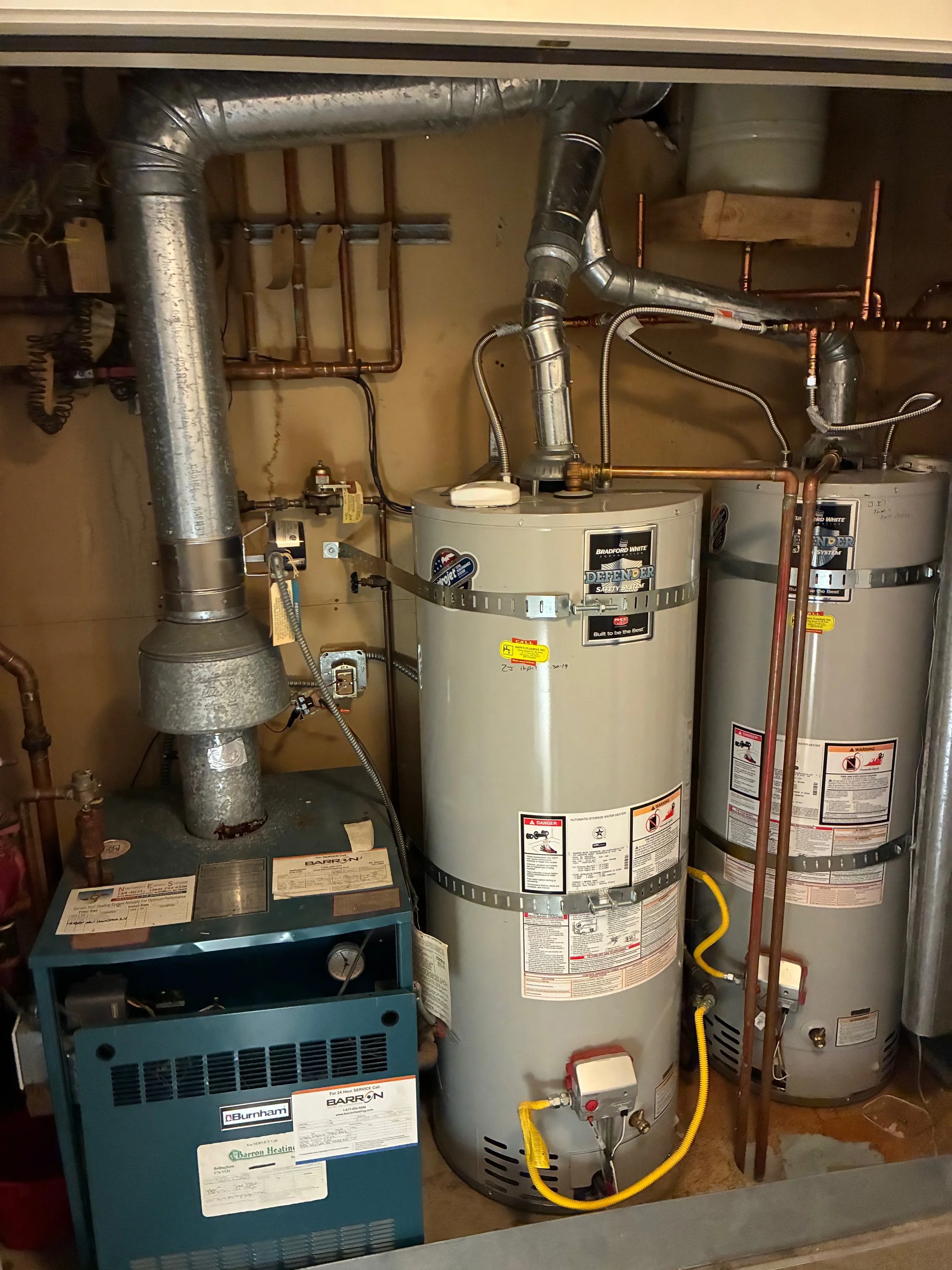 Two water heaters, one gray and one beige, with pipes and a blue heating unit in a utility room.