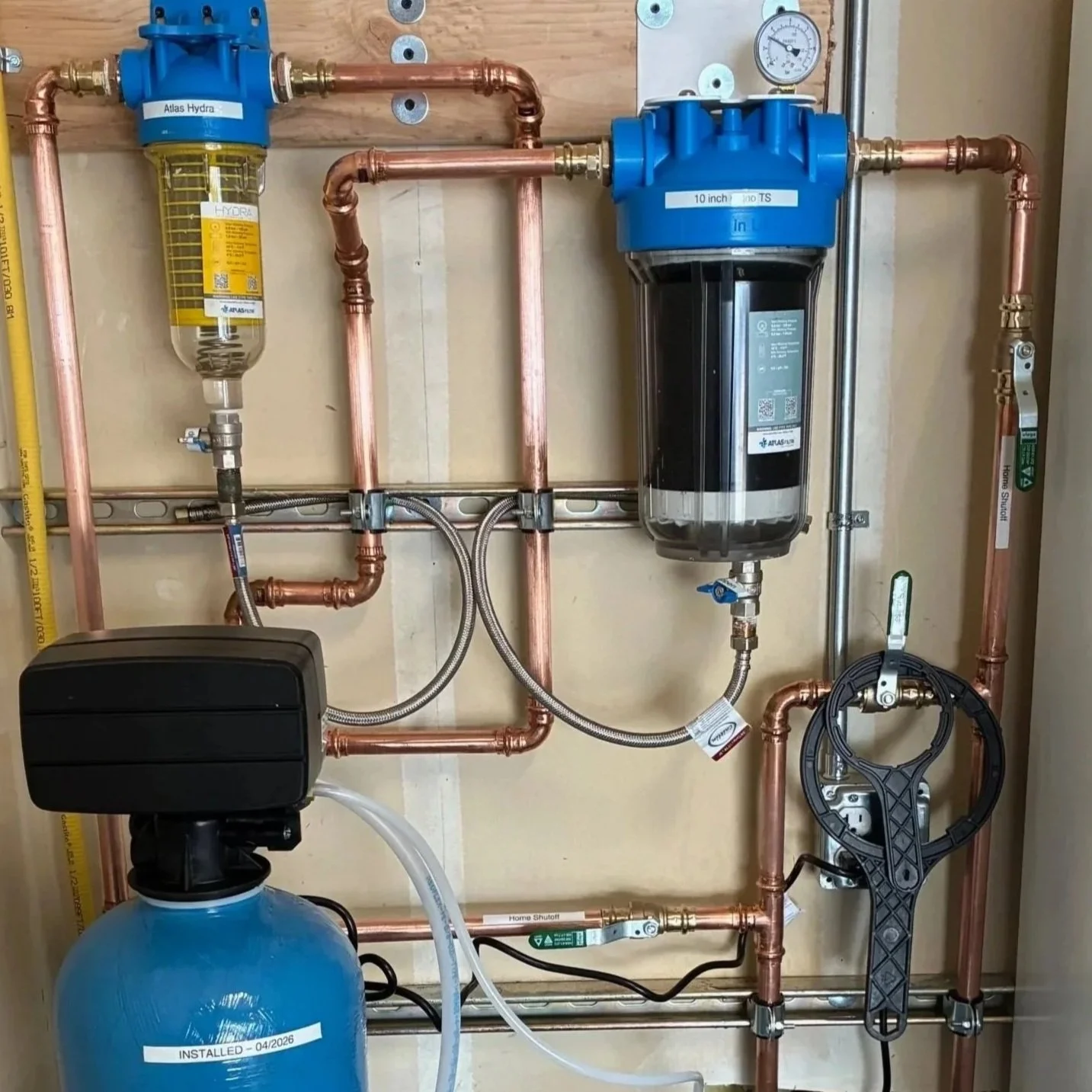 A water treatment system with copper pipes, a blue water filter labeled "Atlas Hydra," a black control valve, a pressure gauge, and a large blue tank labeled "Installed - 04/2026."