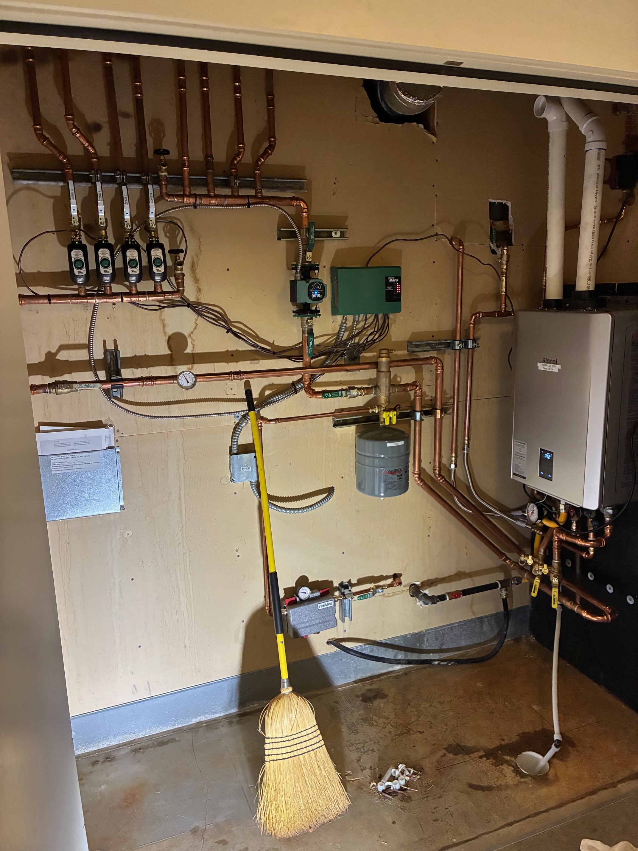 In-progress plumbing installation with copper pipes, gauges, control box, and water heater, in a utility room with unfinished concrete floor and a broom.